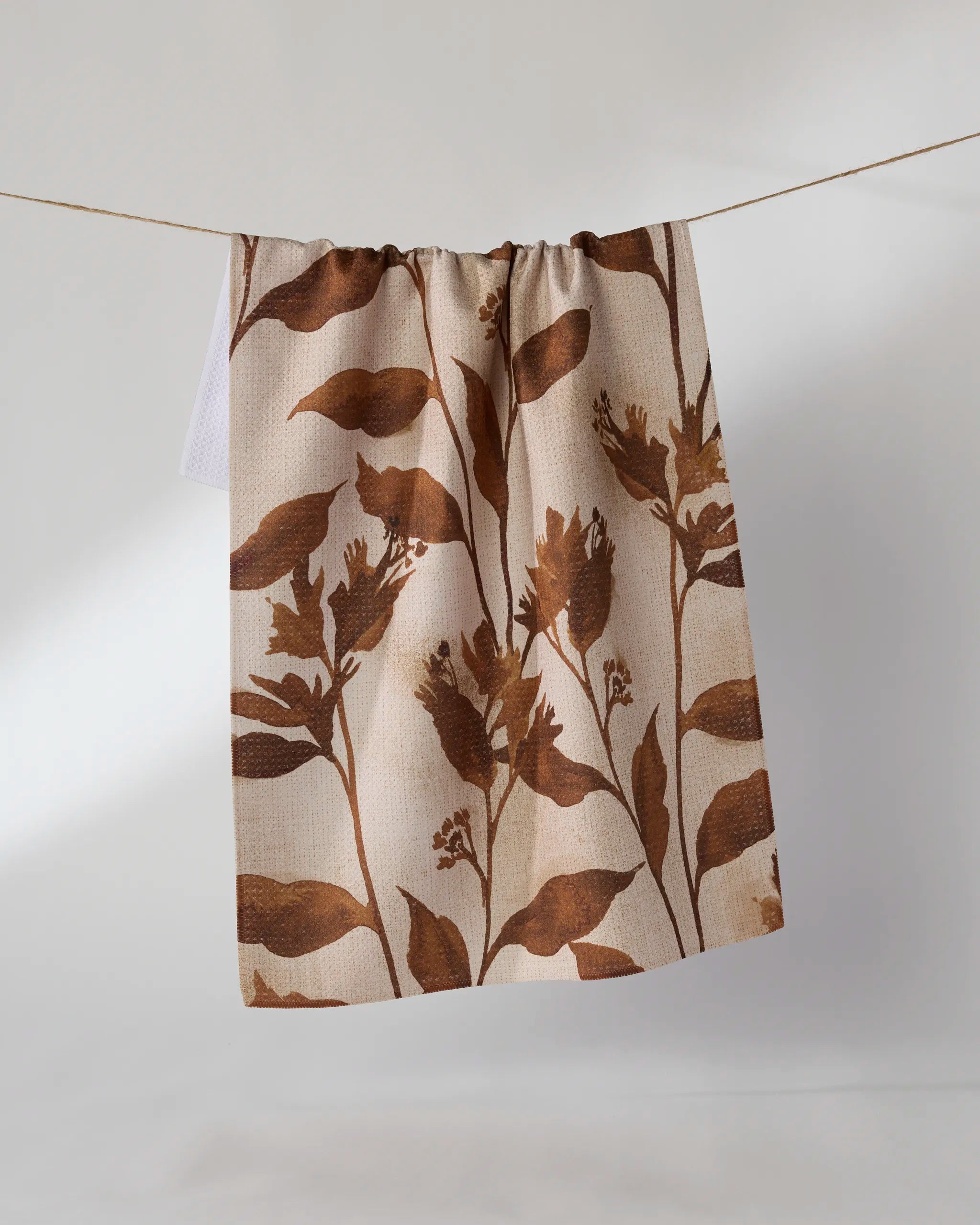 The Willowmere kitchen towel by Geometry, featuring a brown botanical leaf pattern on beige recycled fabric, hangs on a string against a light background.