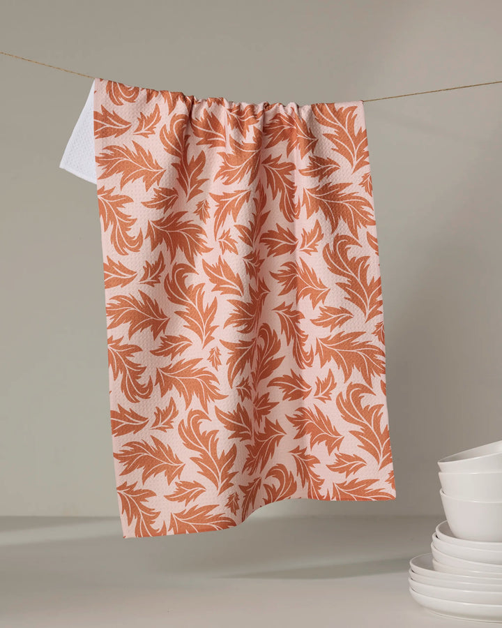 The Wind Fall dish towel by Danelys Sidron, featuring an orange leafy design, hangs against a neutral backdrop. White bowls are stacked in the lower right corner.