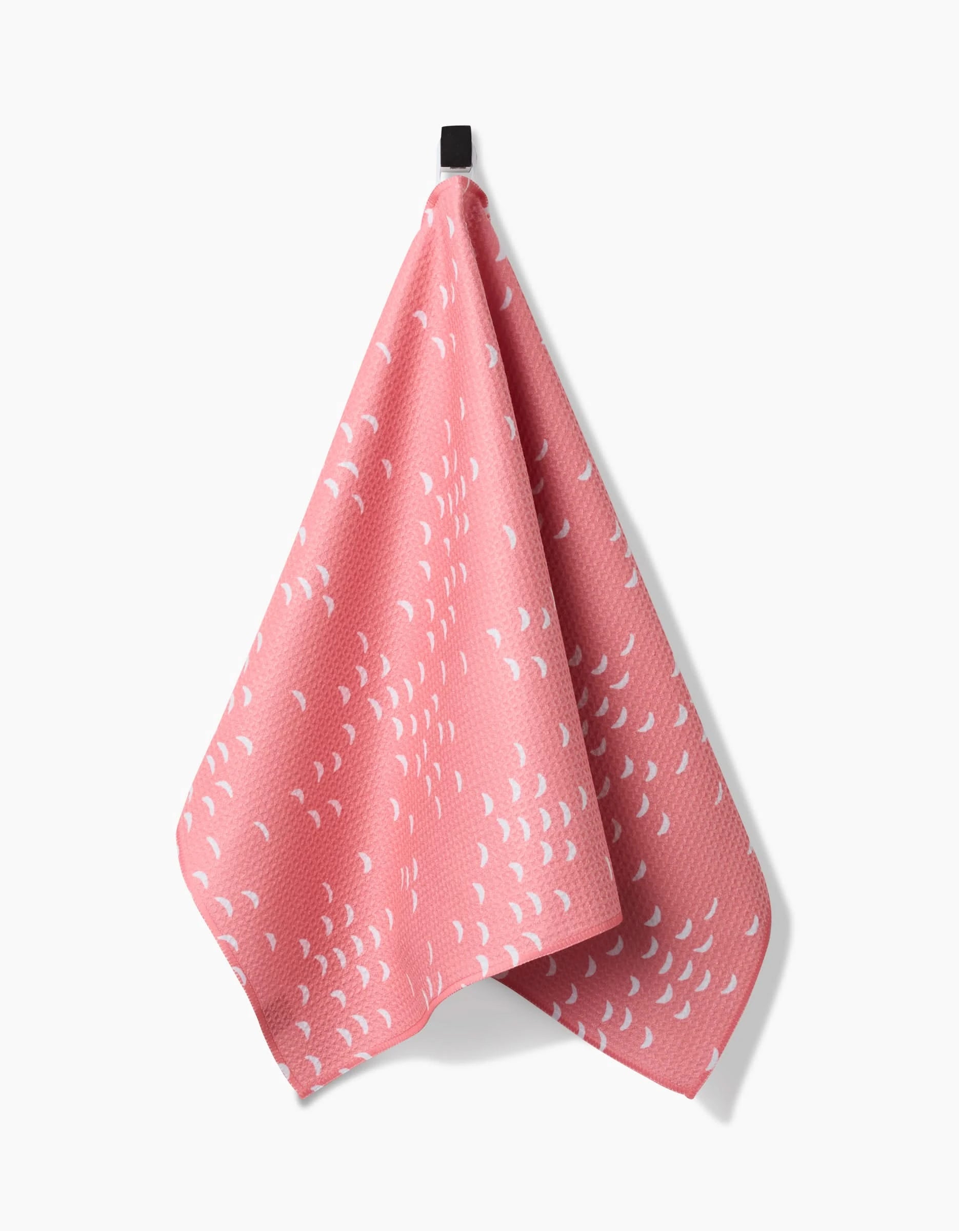 The Jessica Swift Windswept dish towel, featuring a pink textured fabric with a white crescent pattern, hangs from a black hook against a white background.