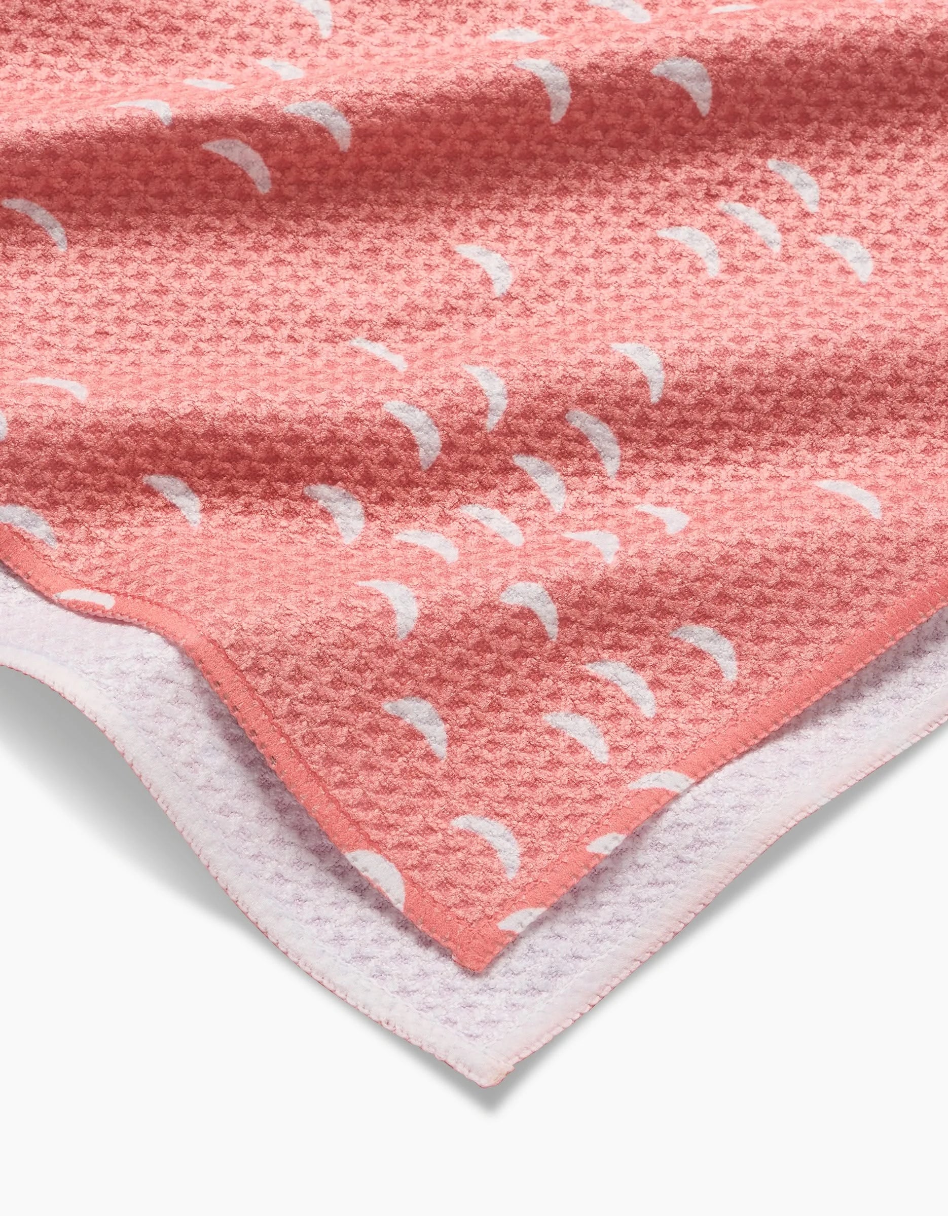 The Windswept blanket by Jessica Swift features a pink textured design with white crescent moons, folded to reveal a soft white underside—ultra-soft and reminiscent of a cozy tea towel.