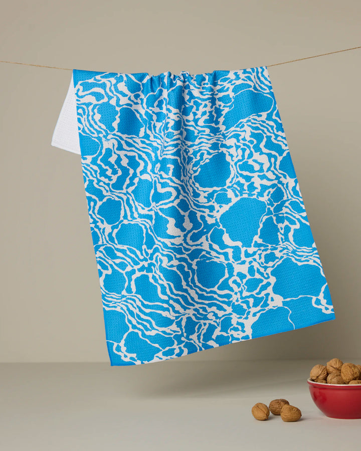 The Geometry Windward kitchen towel, featuring a blue and white abstract wavy design, hangs from a clothesline above a red bowl of walnuts set against a beige background.