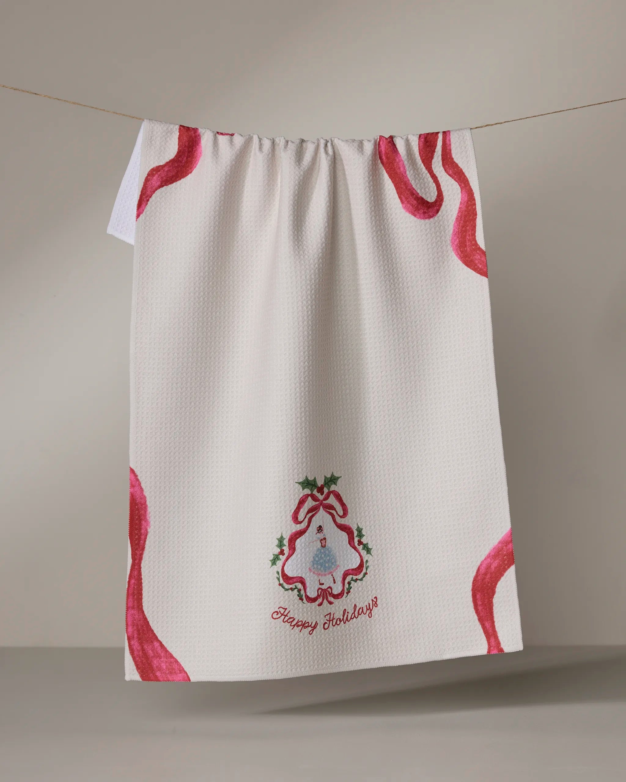 A white quick-drying "Winter Ballerina" tea towel by Geometry hangs on a line, accented with festive red ribbon designs, an embroidered holiday bell with holly, and "Happy Holidays" text at the bottom.