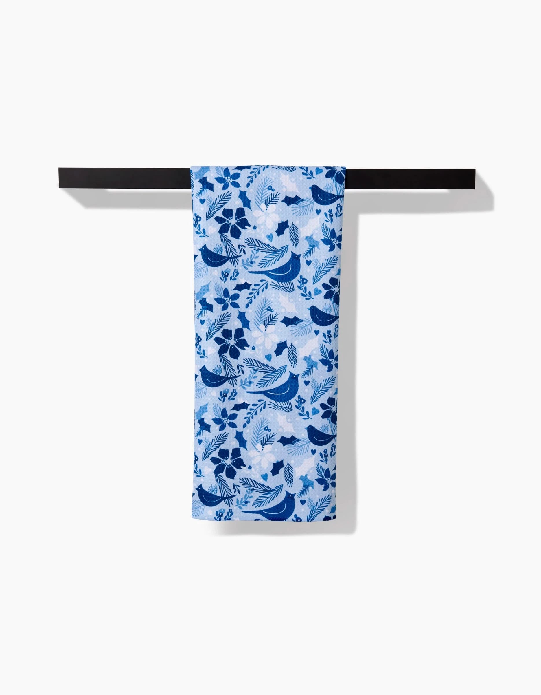 The Winter Birds tea towel by Kris Ruff, decorated with blue and white bird and flower patterns, hangs neatly on a horizontal black bar against a plain white background.