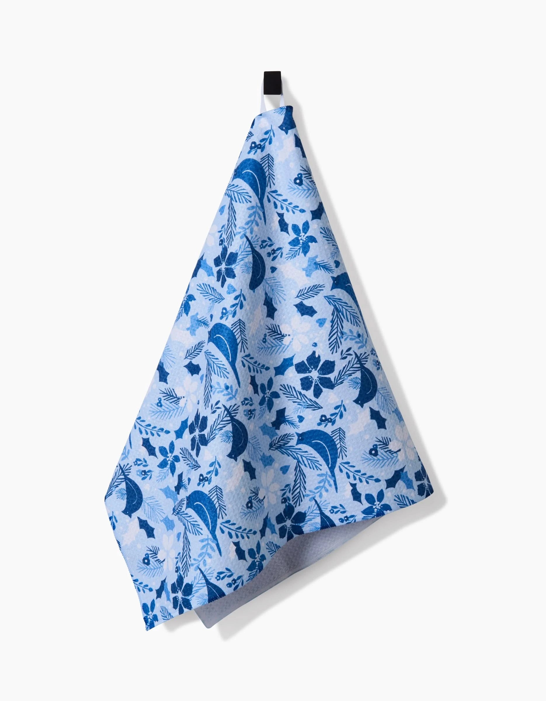 The Kris Ruff Winter Birds tea towel, featuring a blue and white floral pattern and made from ultra-absorbent recycled materials, hangs by a black loop against a plain white background.