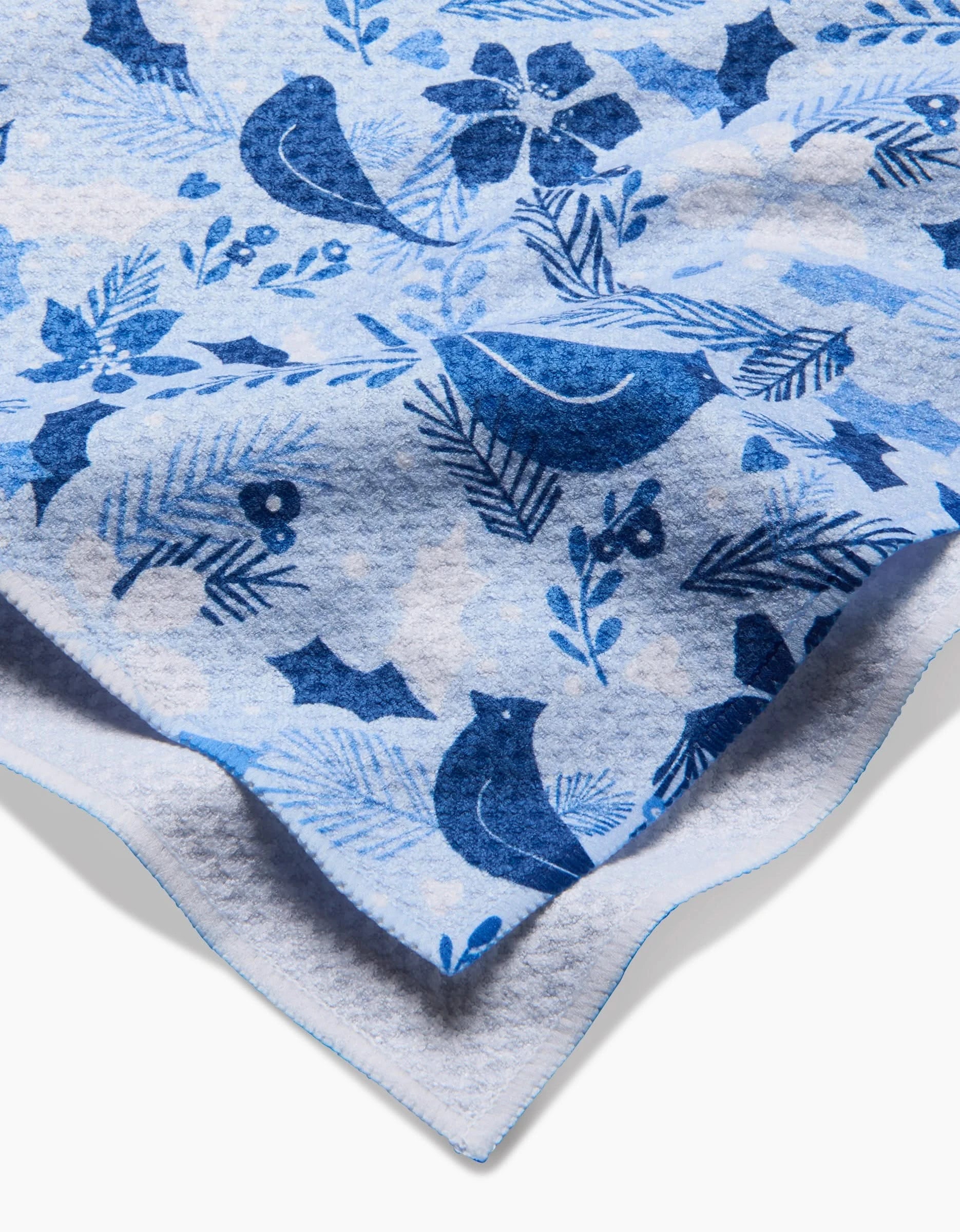 The Winter Birds tea towel by Kris Ruff features a soft, light-blue design with dark blue birds, pine branches, holly leaves, and flowers. Blue-stitched edges and ultra-absorbent, fleece-like fabric add a cozy touch.