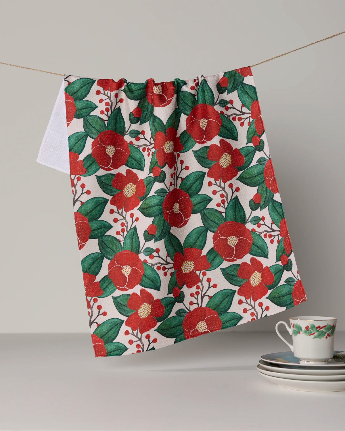 The Geometry “Winter Camellia” kitchen tea towel, featuring vibrant red and green camellia flower patterns, hangs on a string beside a small stack of plates and a teacup adorned with the same floral motif. Set against a neutral background, this ultra-absorbent towel is crafted from recycled materials, bringing both sustainable charm and elegance to your kitchen décor.