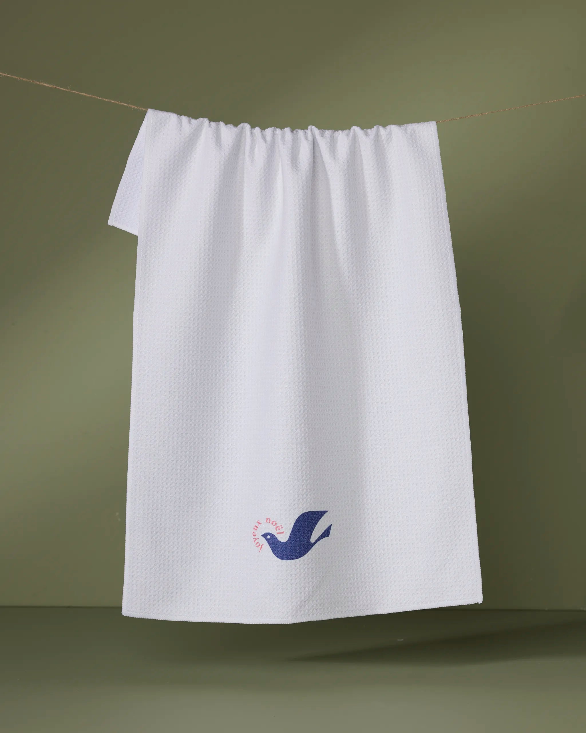 The Geometry "Winter Dove" tea towel, made from recycled materials, features a small blue bird and red text in the corner, hanging on a clothesline against a muted green background.
