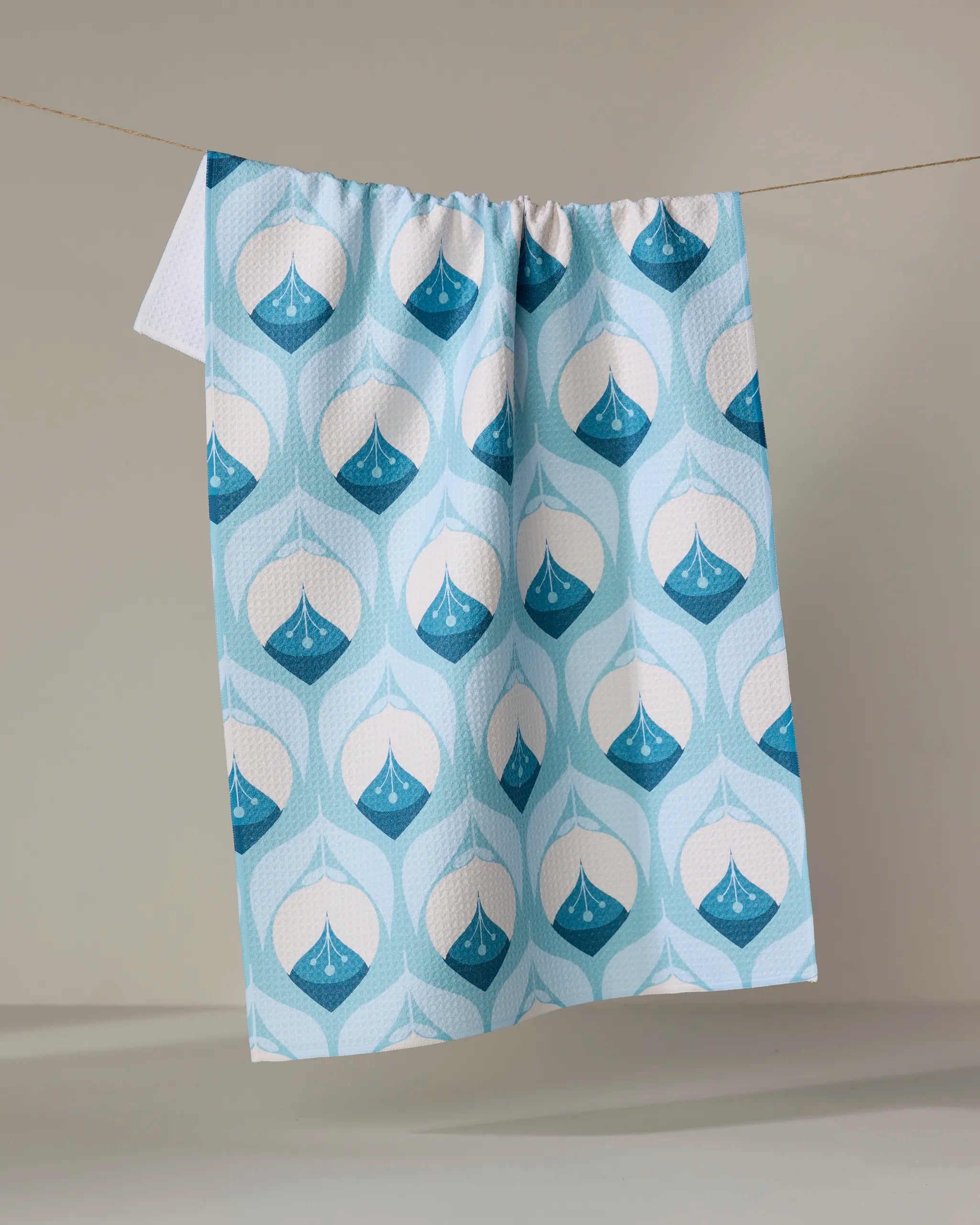 The Blissful Flamingo Winter Florals recycled kitchen towel, featuring a blue and white geometric droplet pattern, hangs on a line against a neutral background.