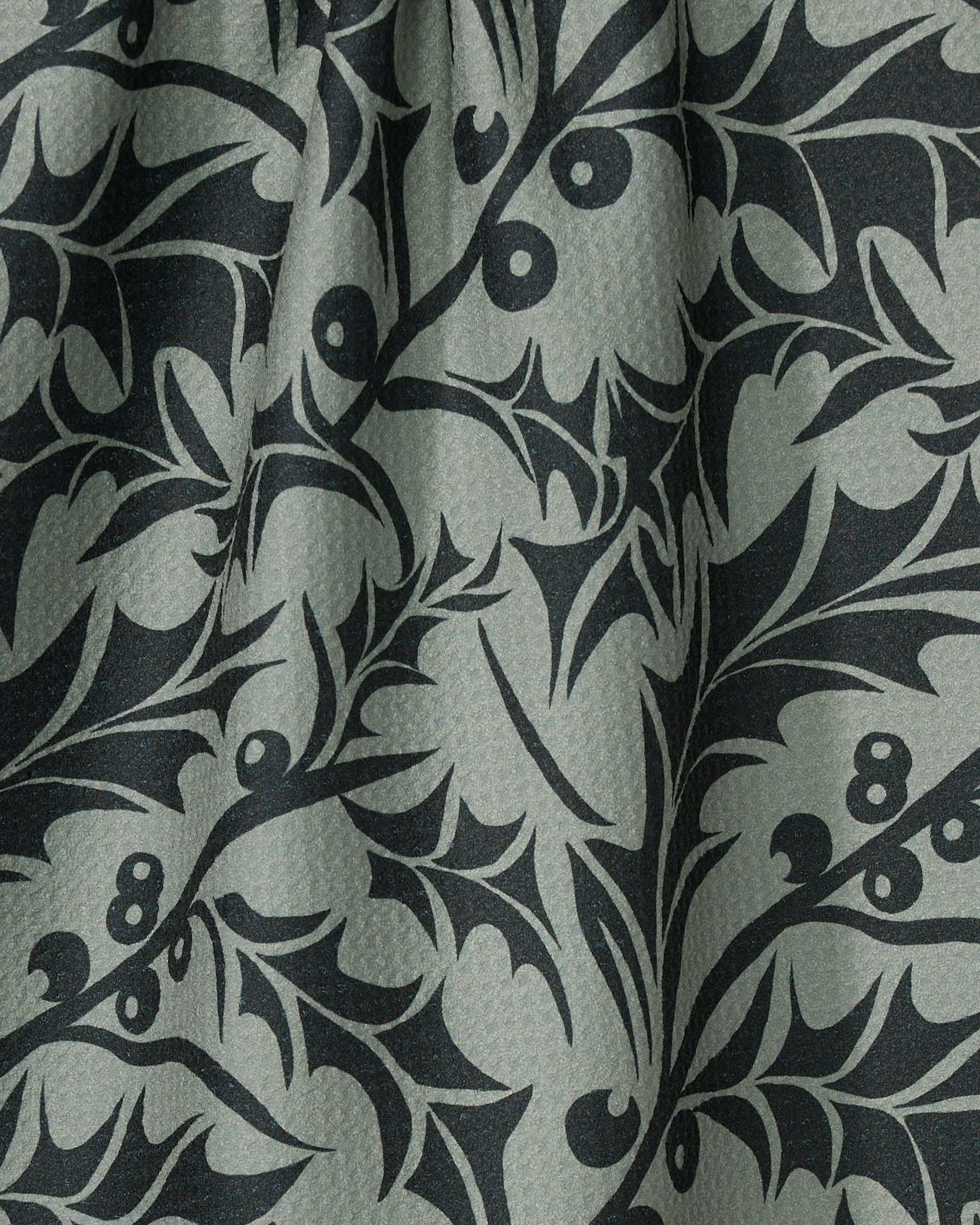 Close-up of Geometry's Winter Foliage kitchen towel, featuring a repeating dark green holly leaf and berry pattern on a lighter green background. The eco-friendly material has a subtly textured surface with soft folds.