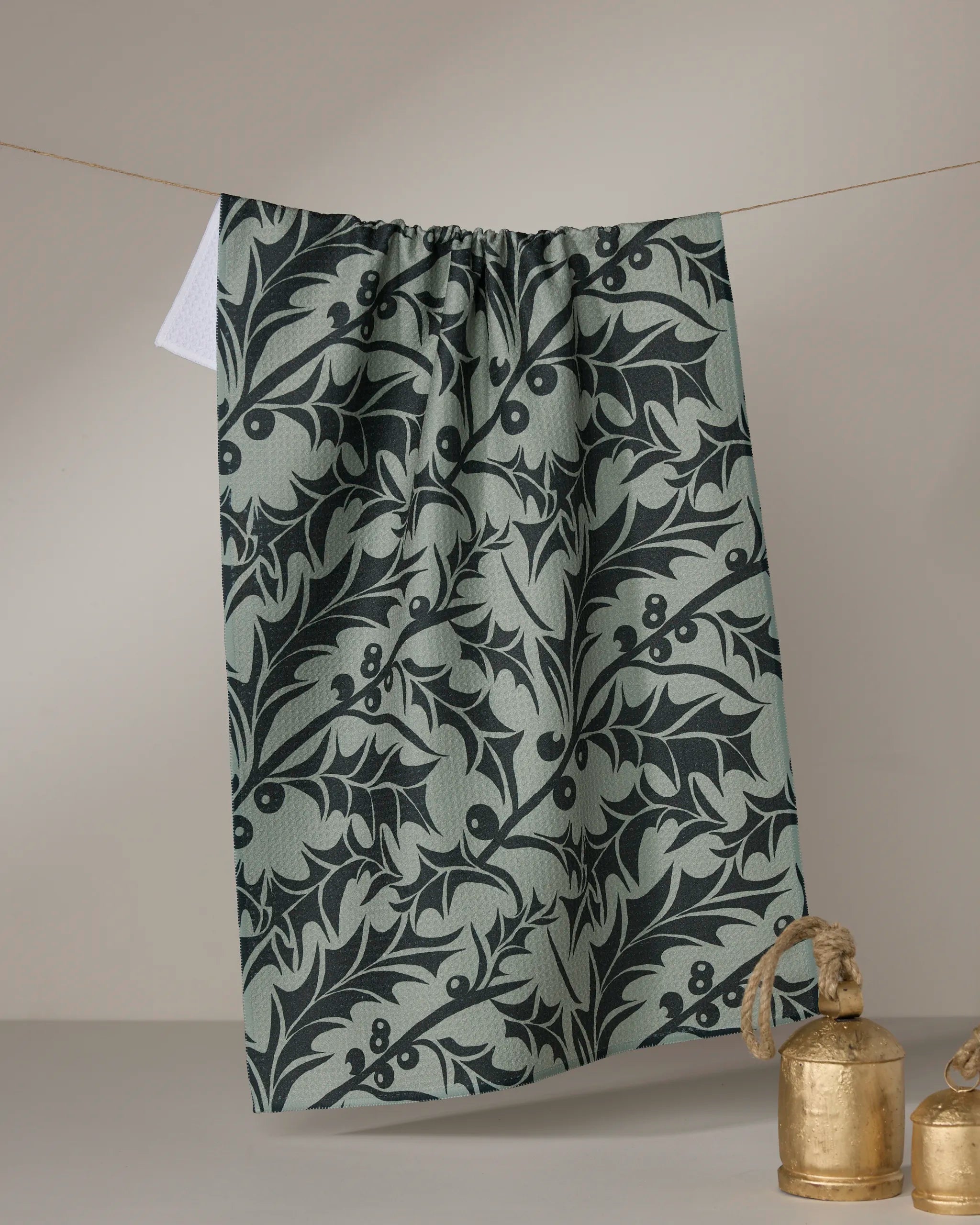 The Winter Foliage tea towel by Geometry, featuring a dark green botanical leaf and berry pattern, hangs on a line against a neutral background, while two gold metal bells sit nearby.