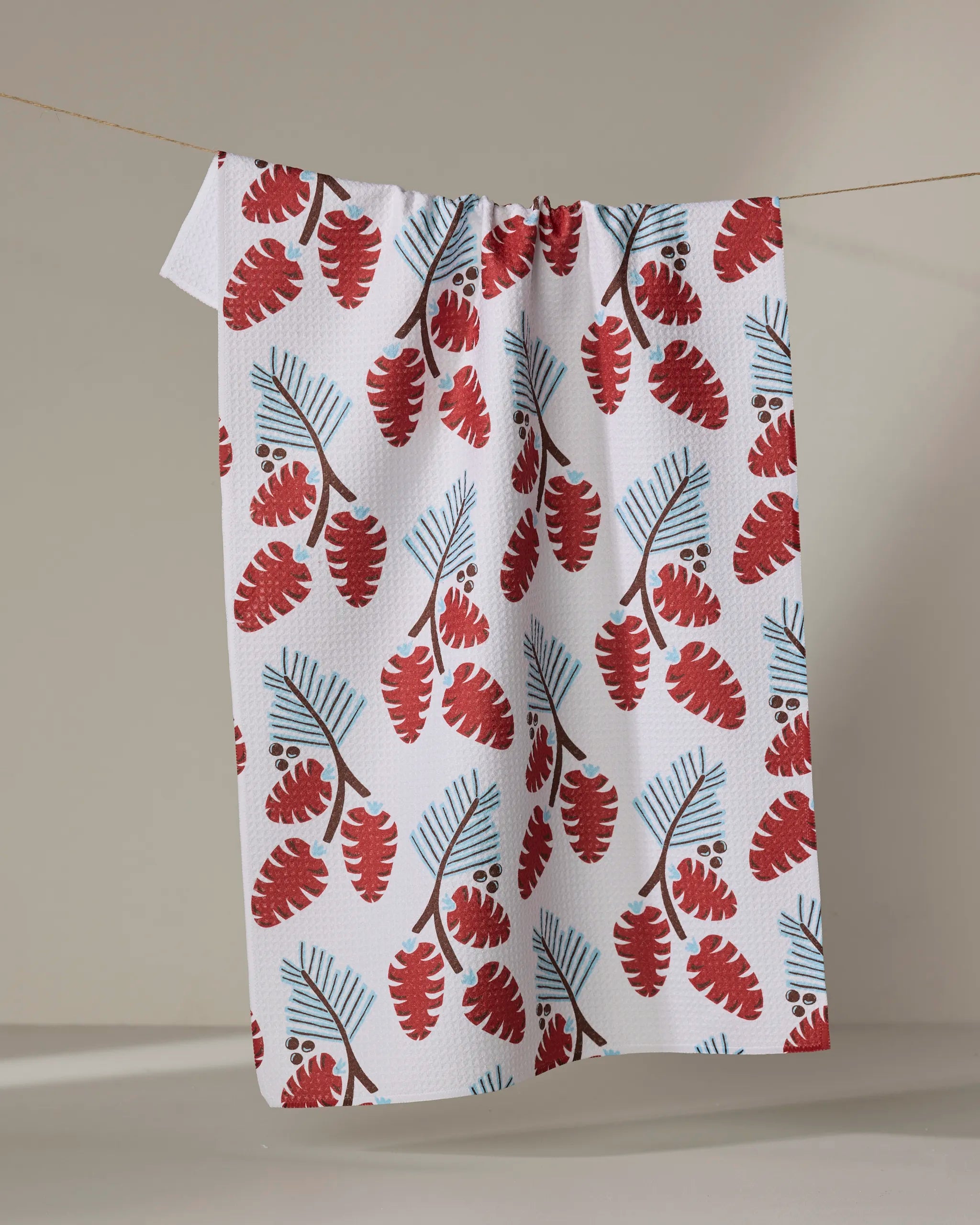 The "Winter Forage" tea towel by Lizzie Clark, featuring red and blue botanical leaves on white recycled fabric, hangs on a line against a plain, neutral background.