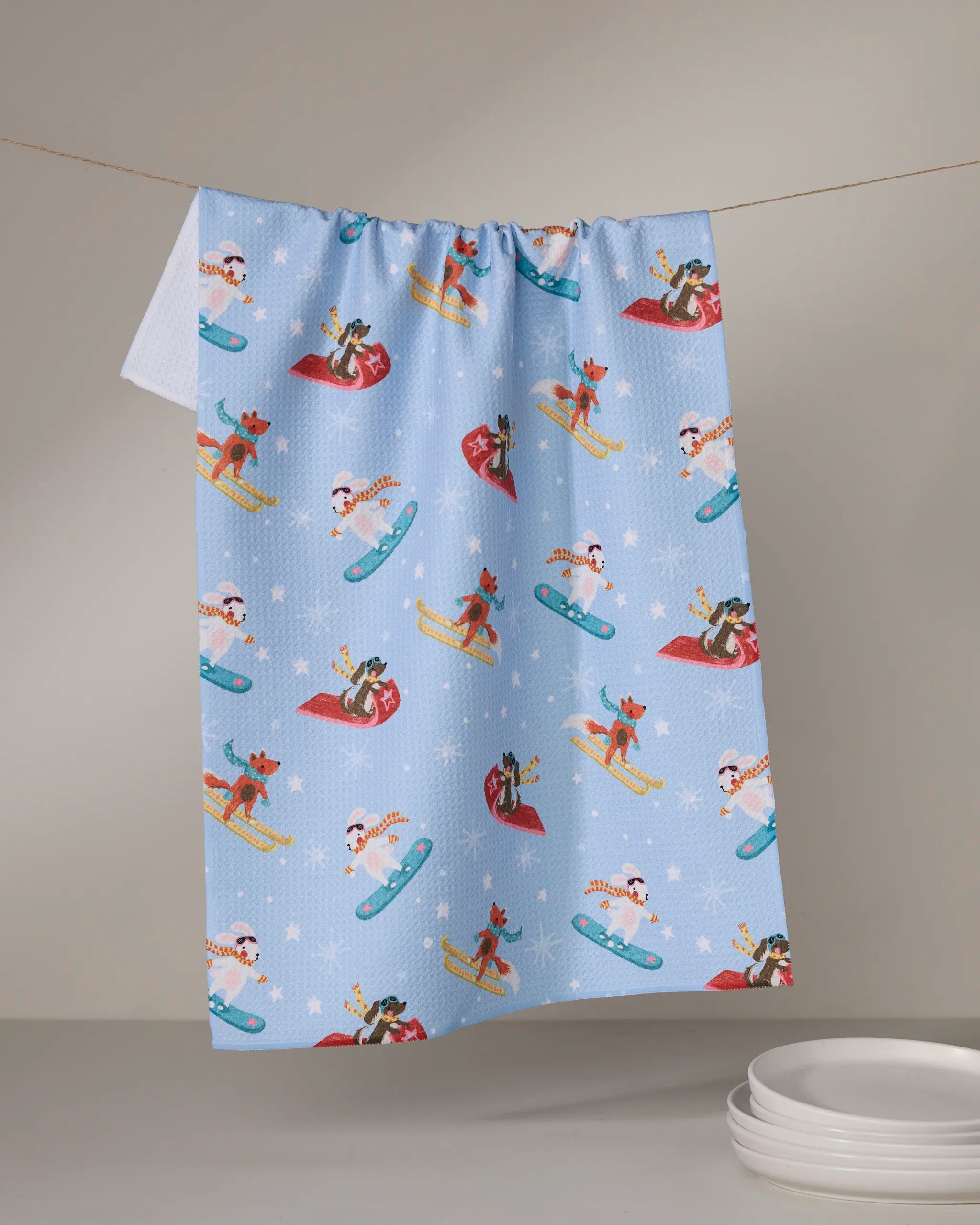 The Geometry Winter Friends tea towel, made from recycled materials and featuring dogs and cats sledding and snowboarding in a light blue winter pattern, hangs on a line above stacked white plates.