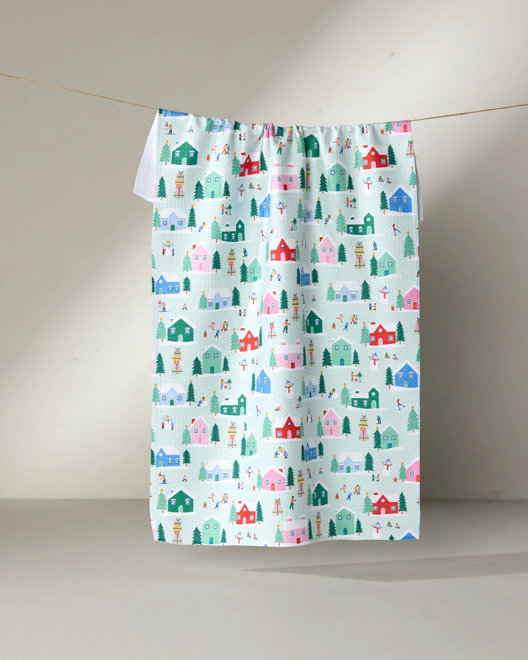 The "Winter Magic" tea towel by Ceyda Alasar is made from recycled materials and displays houses, trees, and snowmen in red, green, pink, and blue on a clothesline against a neutral background.