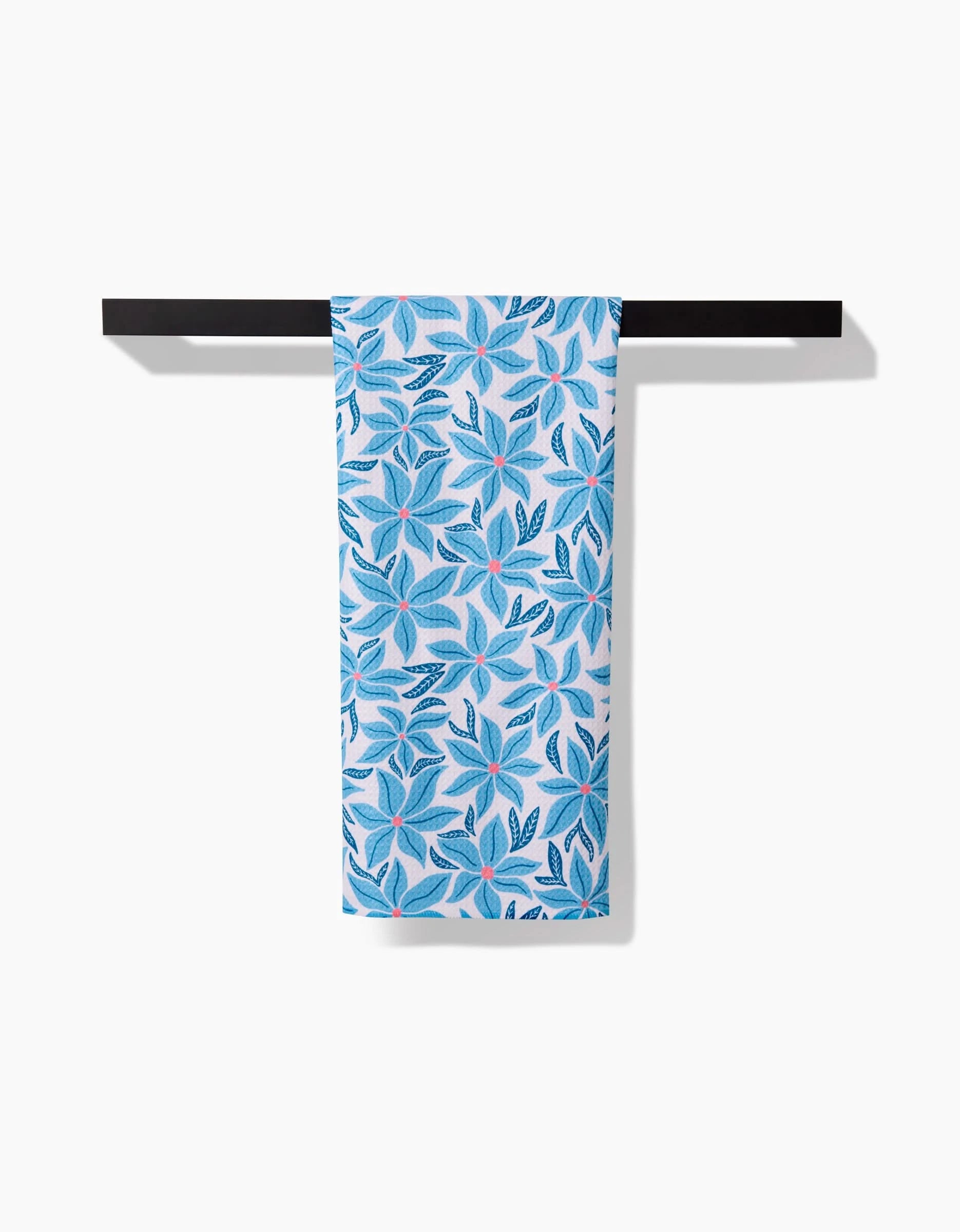 The Kayla Ann Winter Poinsettia eco-friendly kitchen towel, featuring a blue and teal leaf pattern, hangs neatly on a black towel bar against a white background.