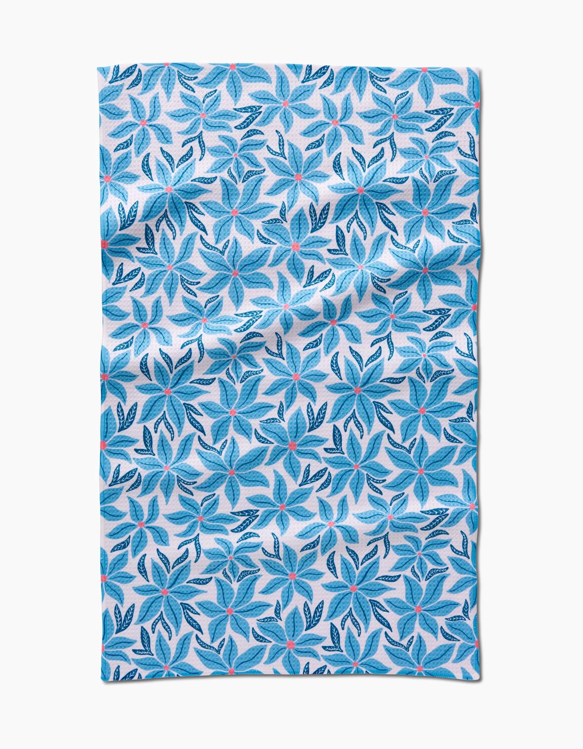 The Kayla Ann Winter Poinsettia tea towel, made from recycled materials, features blue and navy leaf patterns with red dots on a light background and is shown flat with some natural wrinkles.