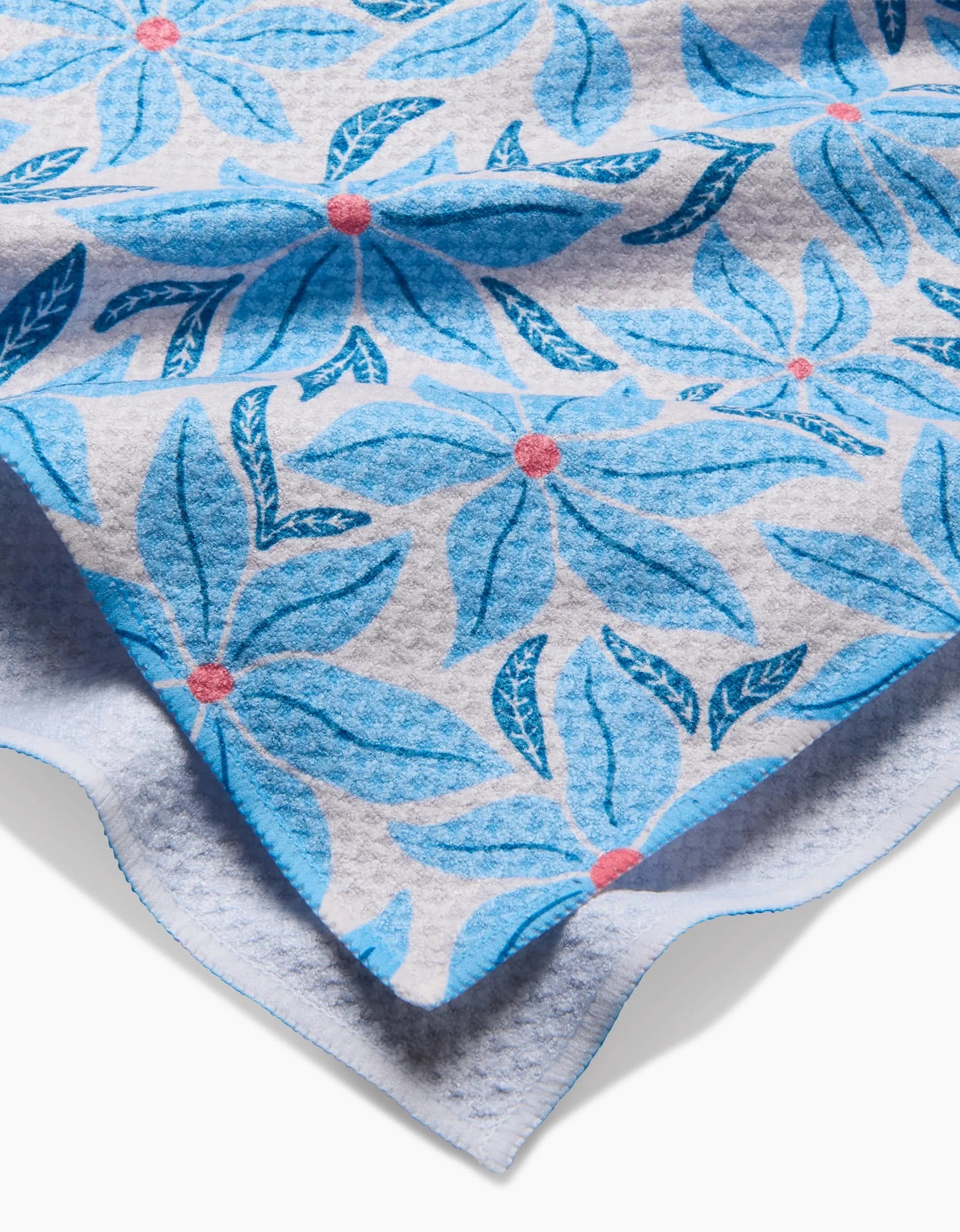A close-up of the Kayla Ann Winter Poinsettia tea towel shows its ultra-absorbent recycled fabric, blue floral pattern with pink centers, large blue leaves on white, and textured edges with a gentle fold highlighting its thickness.