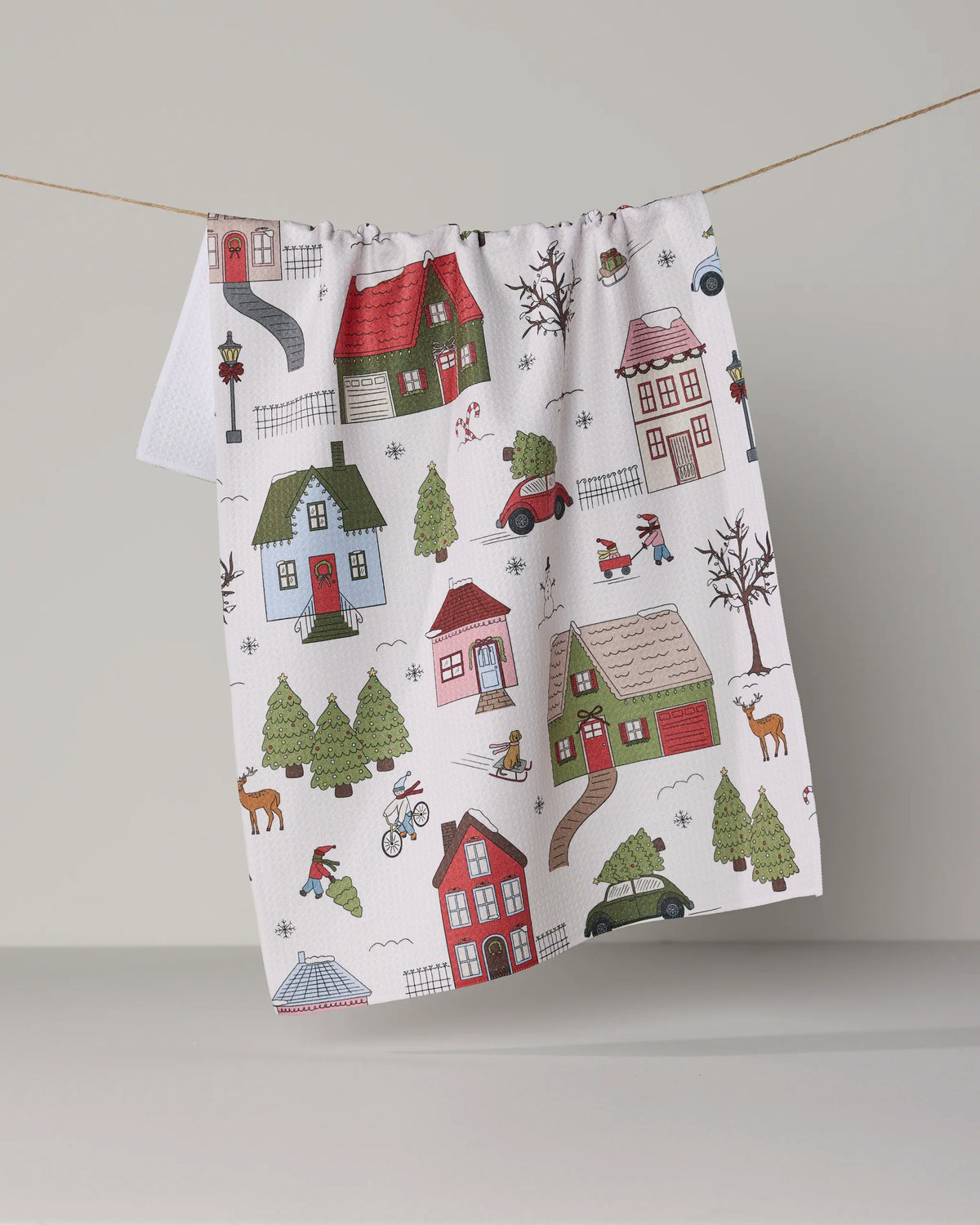 The Geometry “Winter Village” tea towel, made from recycled materials, features a festive village scene with colorful houses, trees, cars, people, and animals on a white background and is shown hanging on a clothesline.