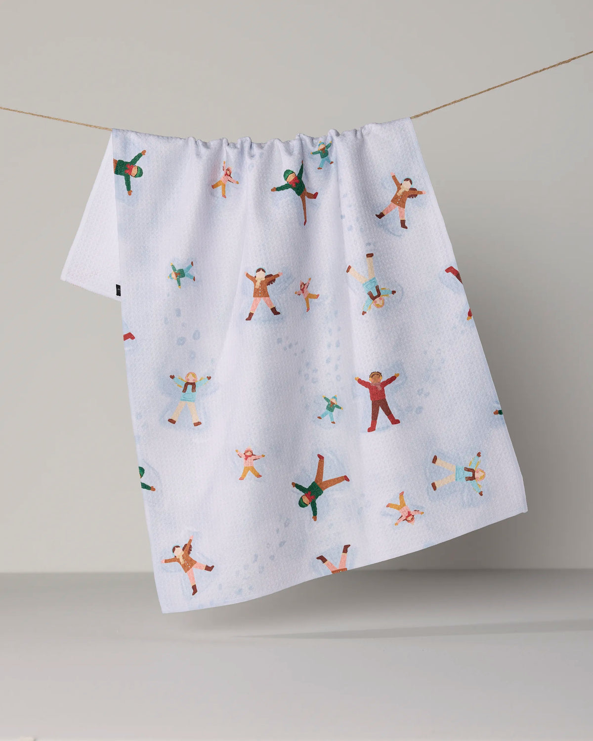 The Geometry “Winter Whimsy” Tea Towel hangs from a string, showcasing a vibrant pattern of small people making snow angels across its white surface. Made from innovative Geoweave™ fabric and recycled materials, this towel blends playful design with sustainability.
