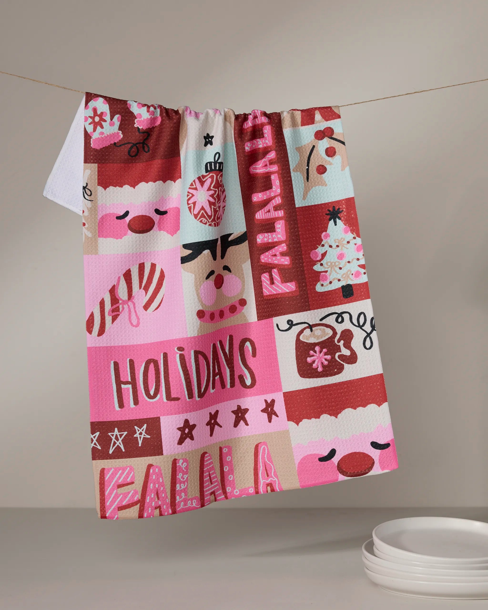 The Winter Wonder kitchen towel by Geometry hangs on a line, featuring festive pink and red holiday illustrations and "HOLIDAYS" and "FALALA." Made from recycled materials. White plates sit nearby.
