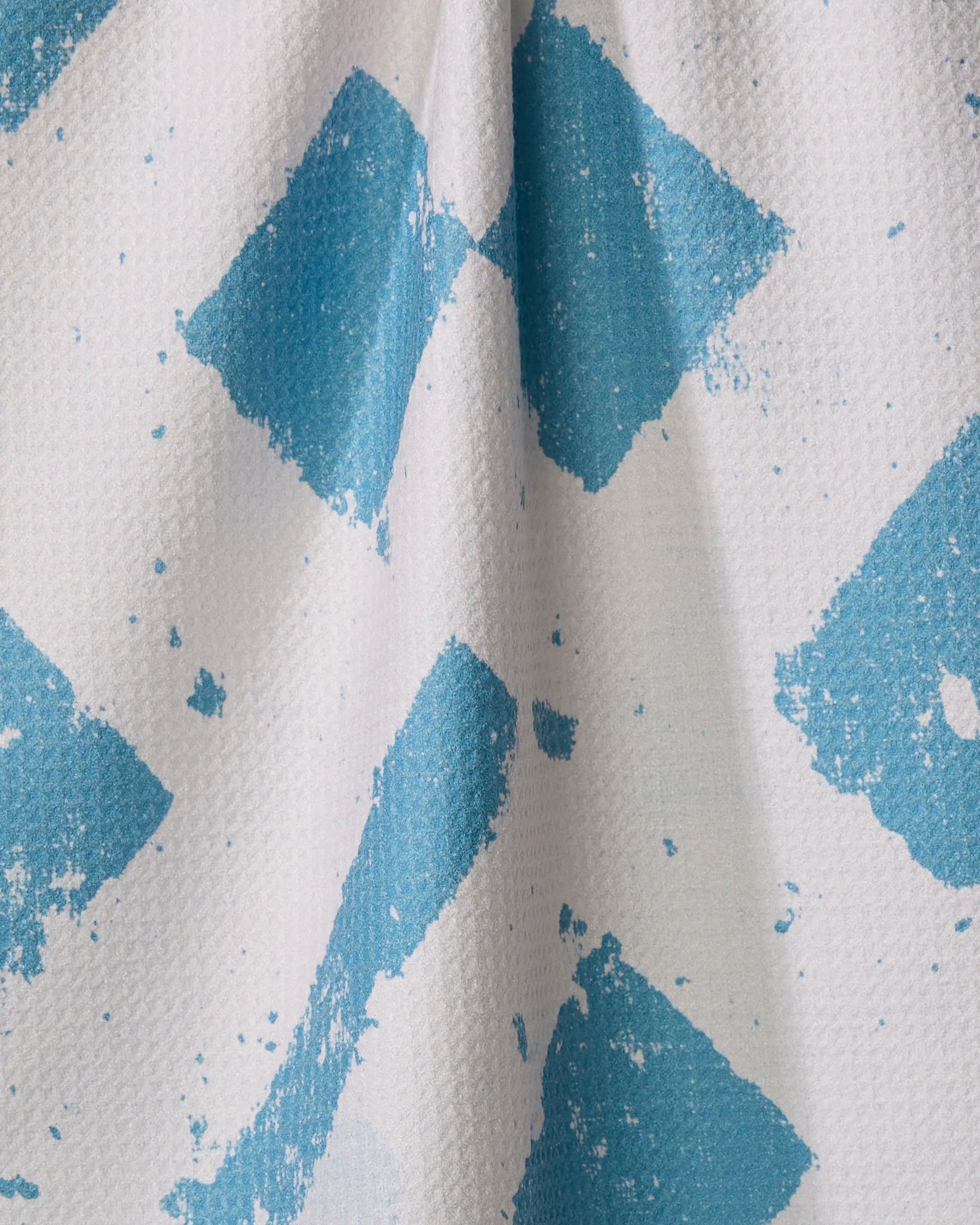 A close-up of the Geometry Wisp Blue tea towel, crafted from recycled materials with a white textured surface, blue diamond patterns, and paint splatters. Draped fabric forms gentle folds and soft shadows.