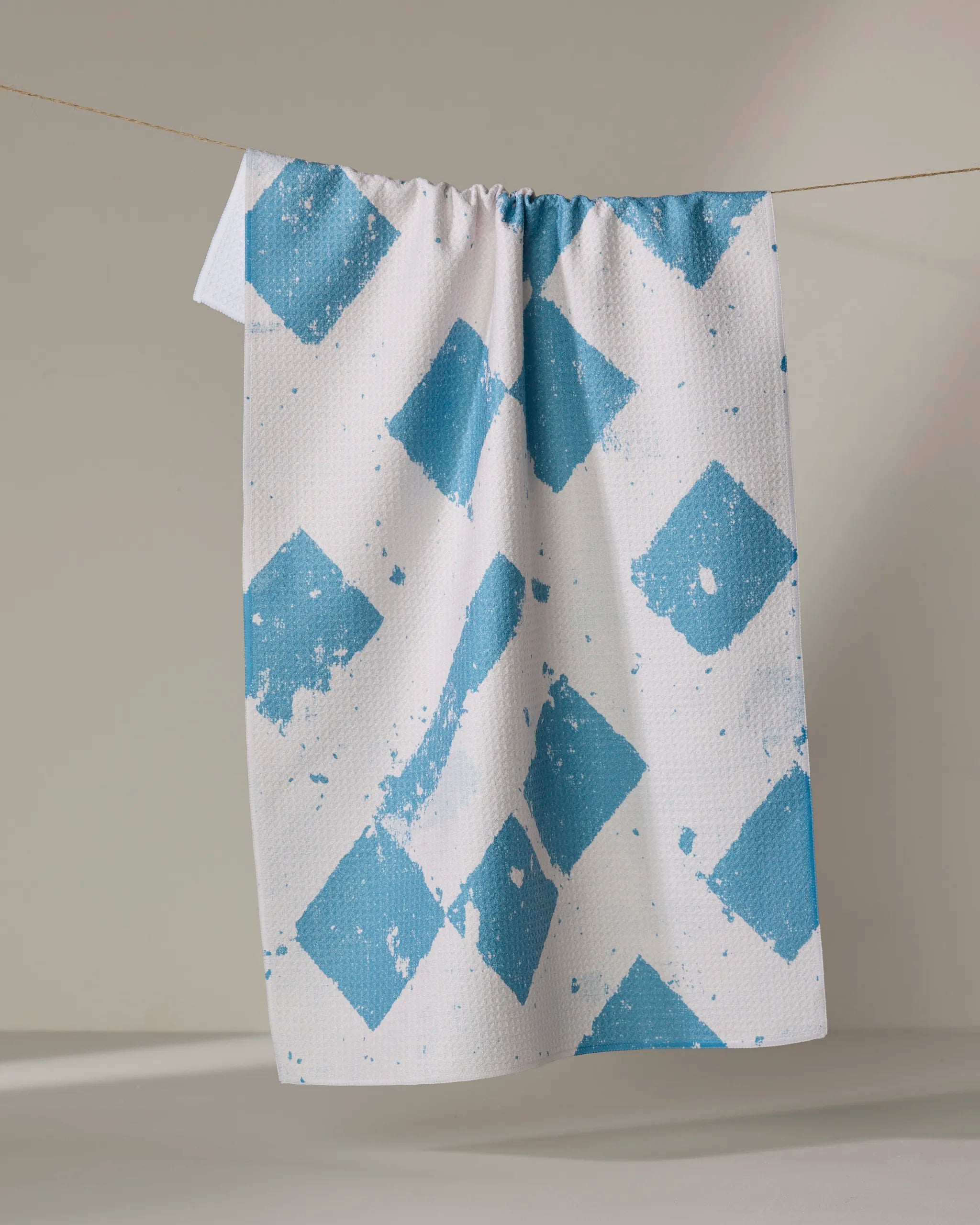 The Geometry Wisp Blue tea towel, made from recycled materials, features a textured blue diamond pattern on white. It hangs on a clothesline against a neutral background, with the diamonds showing a subtle splattered effect.