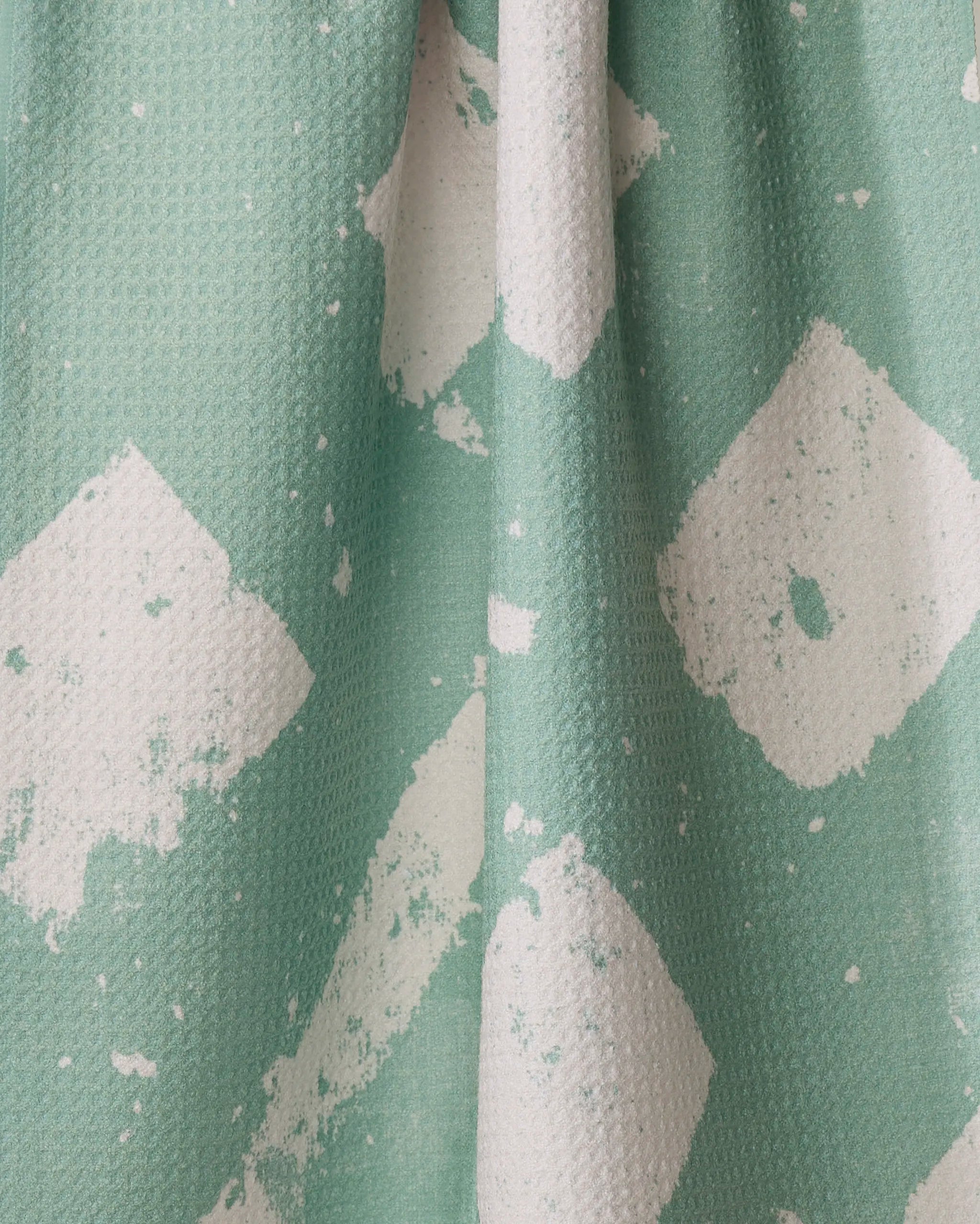 A close-up of Geometry’s Wisp Green kitchen towel shows its eco-friendly, textured fabric with abstract white paint splatters and diamond shapes. Soft folds and shadows highlight this tea towel’s unique design.