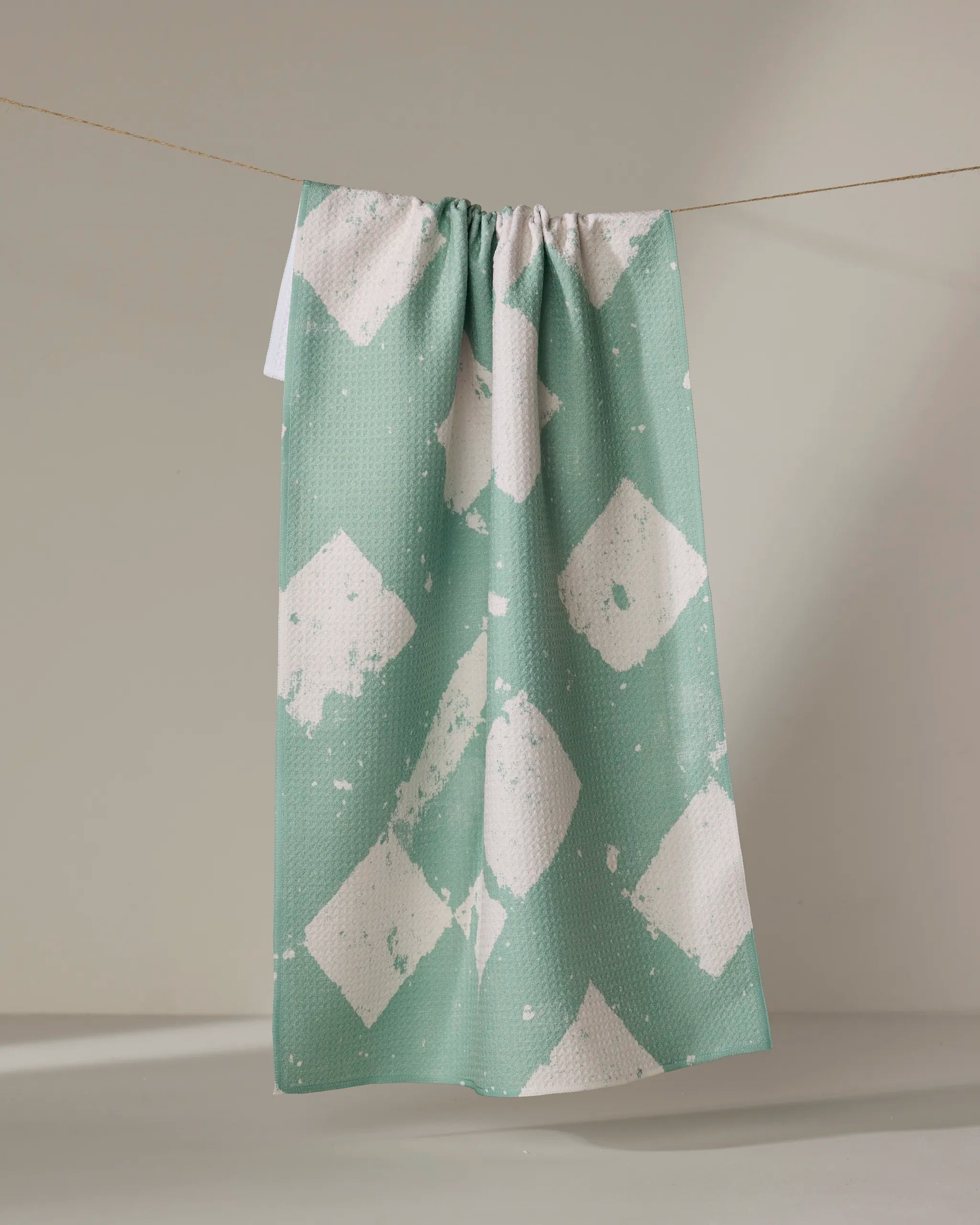 The Geometry Wisp Green kitchen towel, featuring a pale green color with a white diamond pattern, hangs folded over a thin clothesline against a light gray background.