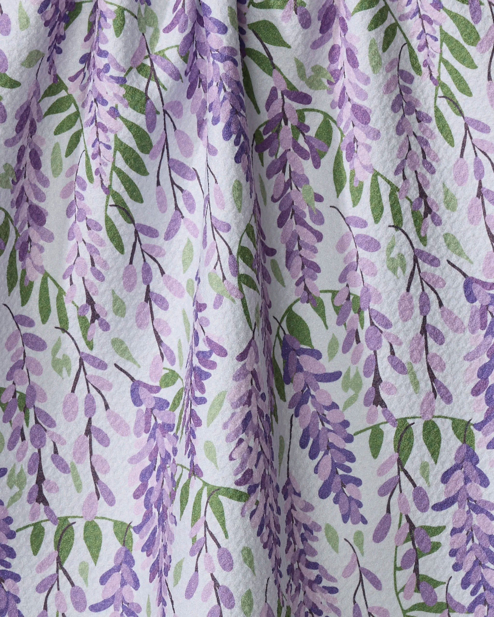 A close-up of Rebecca Bobko's Wisteria fabric shows a light background adorned with purple wisteria flowers and green leaves. Made from recycled materials, its gentle folds make it ideal for a charming tea towel.