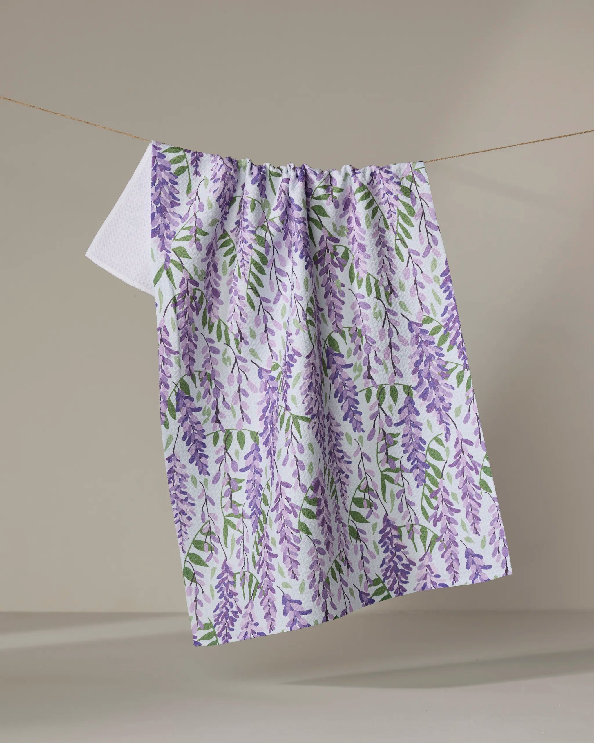 The Rebecca Bobko "Wisteria" kitchen towel, featuring a purple and green floral pattern, hangs on a clothesline against a plain beige background.