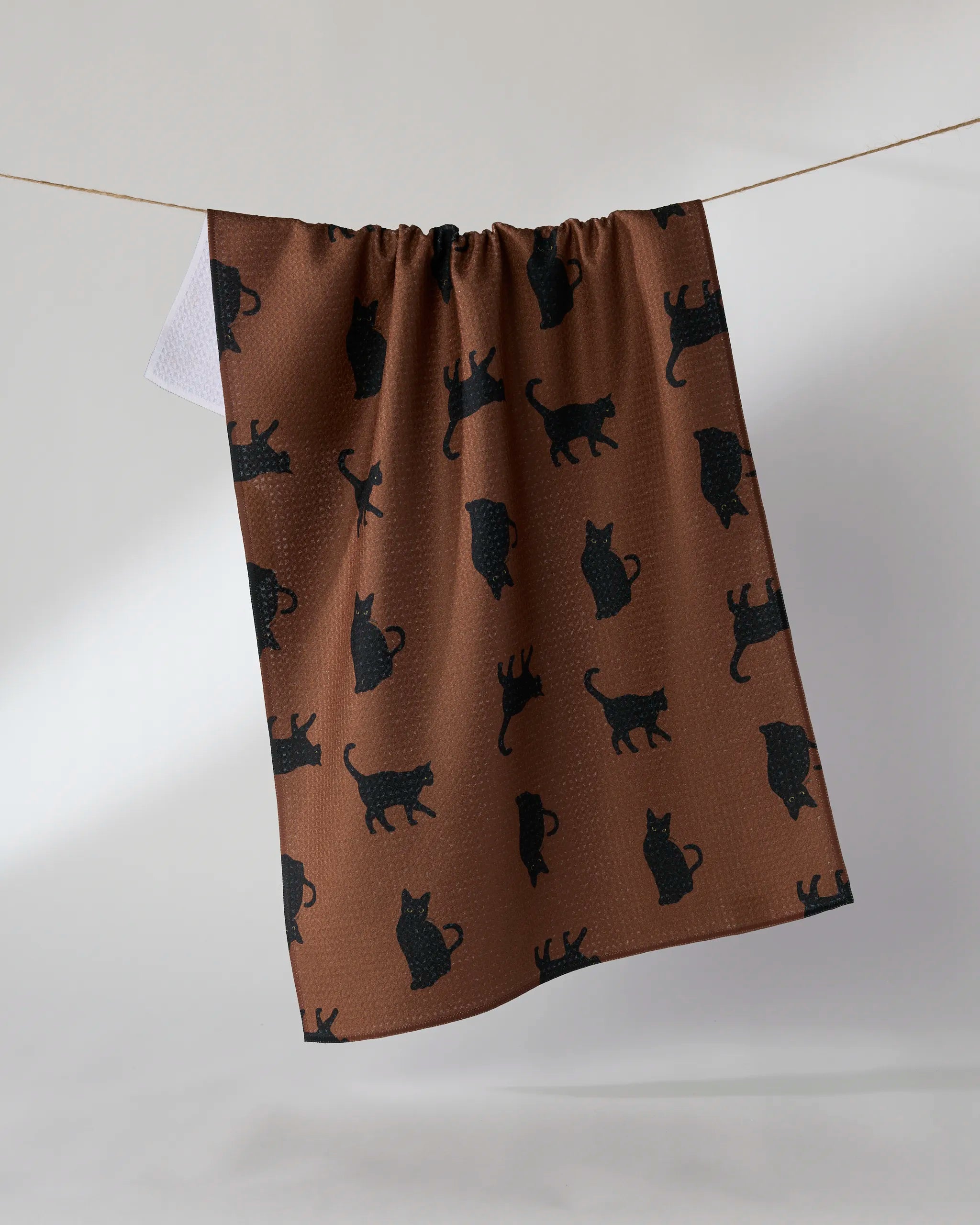 The Geometry Witch's Cat tea towel, featuring black cat silhouettes on brown and made from recycled materials, hangs on a clothesline against a plain white background.