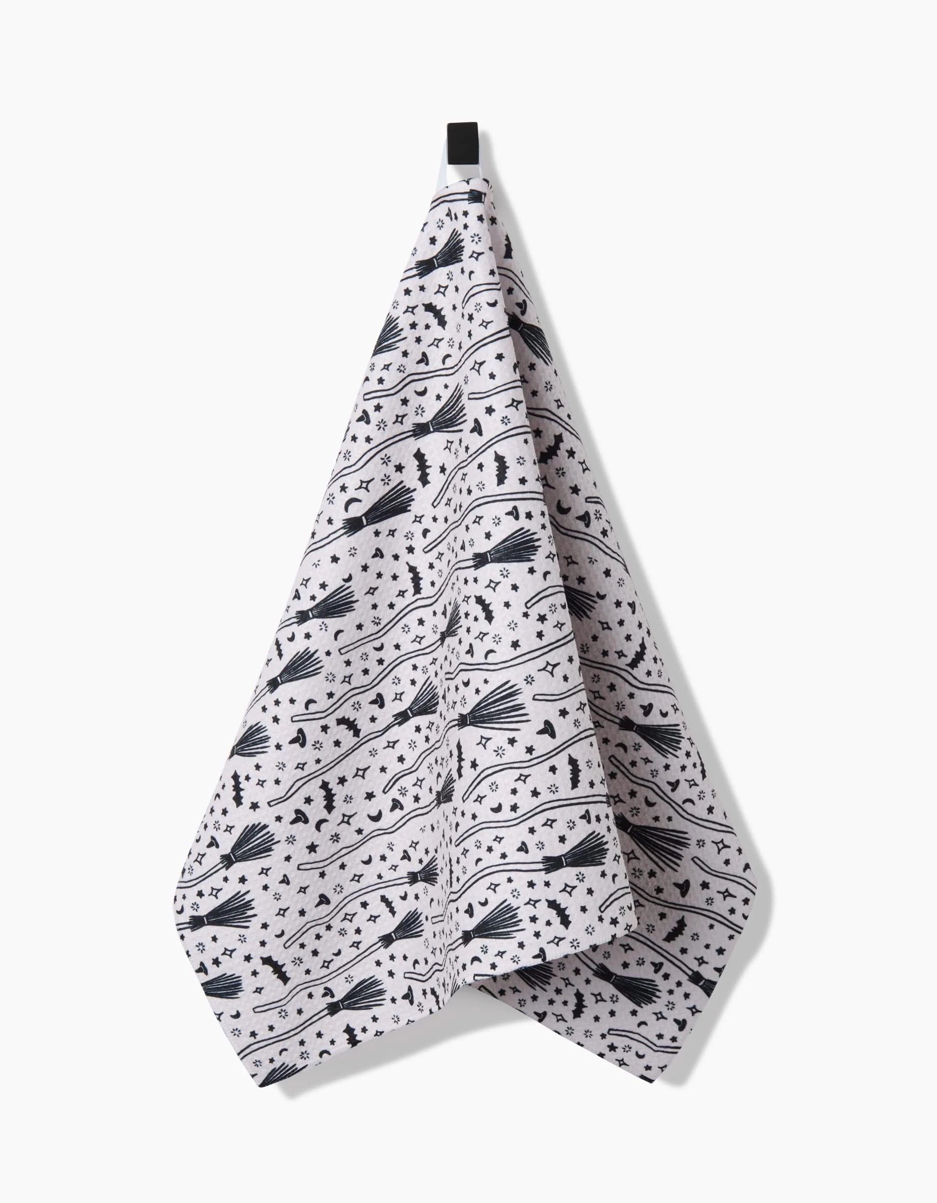 The Kayla Ann Witches Broom tea towel is crafted from recycled materials and features a black witch-themed pattern with flying witches, moons, stars, and broomsticks. It hangs by a single loop against a plain white background.