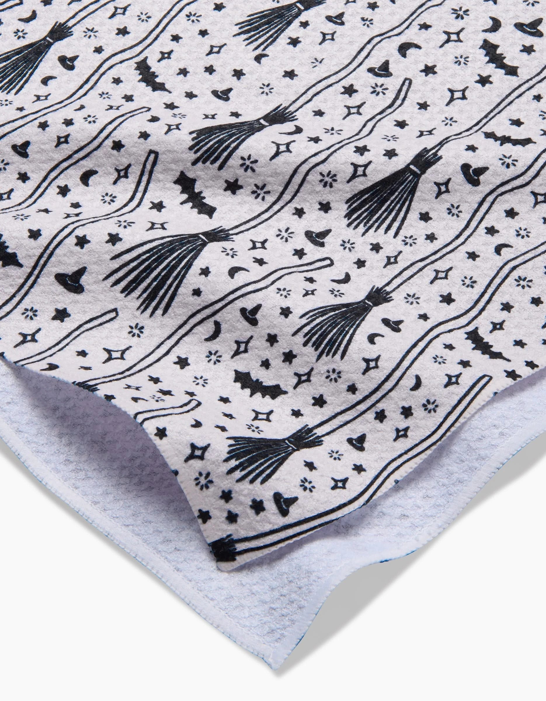 A close-up of the Kayla Ann Witches Broom tea towel, crafted from ultra-absorbent recycled materials, highlights its white fabric decorated with whimsical Halloween witch hats, brooms, bats, stars, and moons.