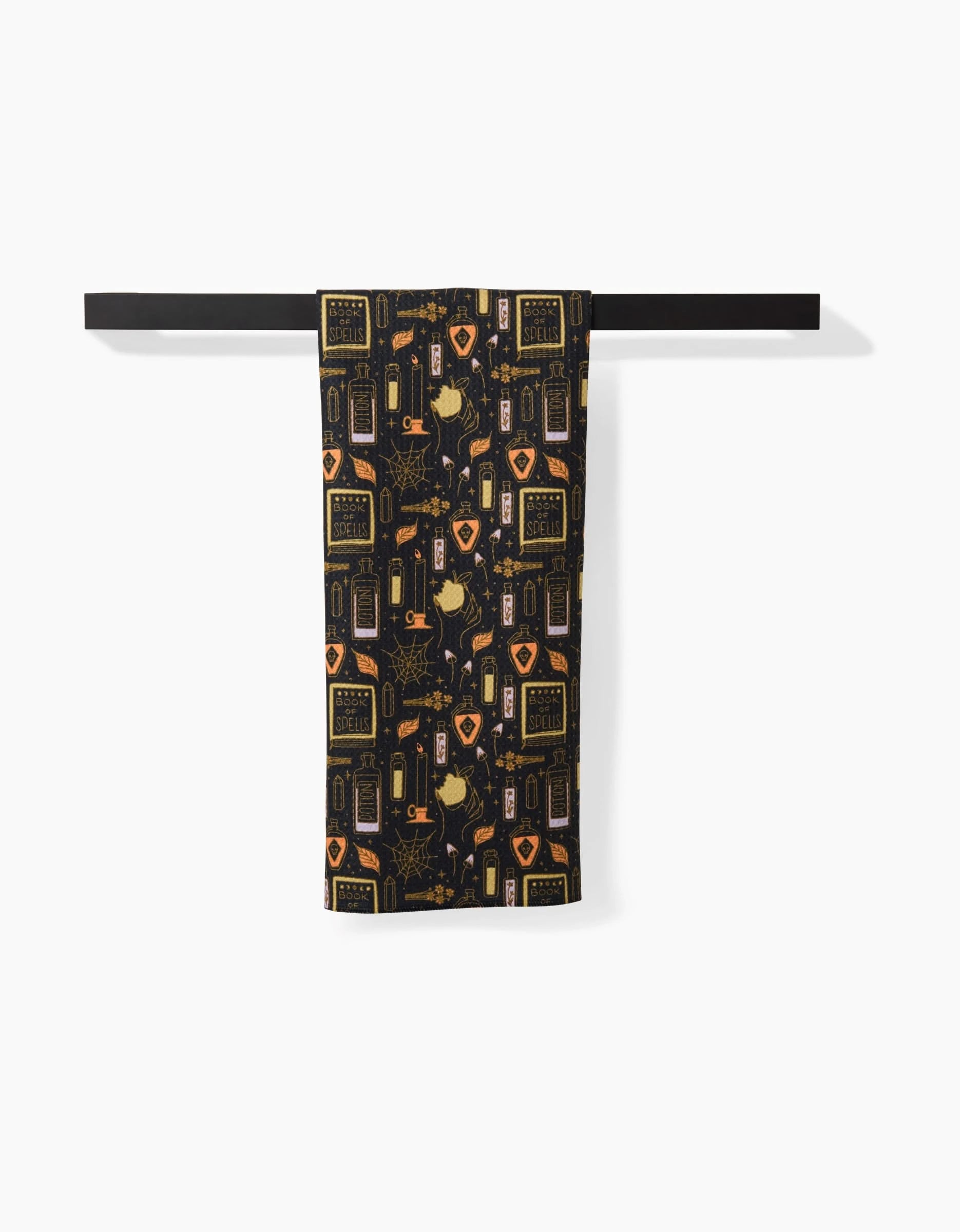 The Mandy Arroyo Witchy Things black tea towel features yellow and orange potion bottles, labels, plants, and mystical symbols. Made from recycled materials, it’s eco-friendly and displayed hanging on a black rod against a white background.