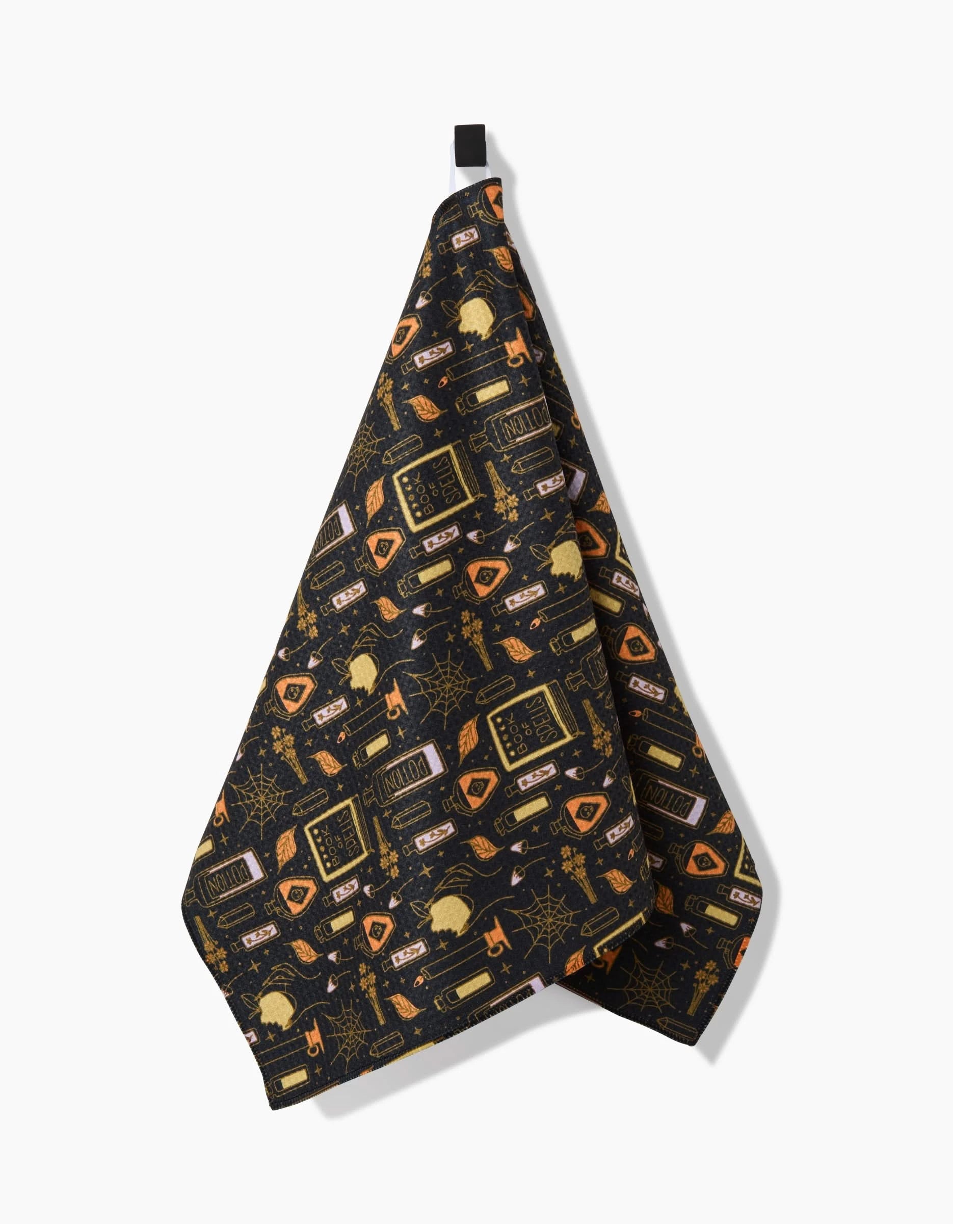 The Witchy Things kitchen towel by Mandy Arroyo features black, absorbent fabric with orange and gold Halloween motifs—spider webs, cauldrons, potion bottles, black cats, and skulls. Made from recycled materials and shown hanging against a white background.
