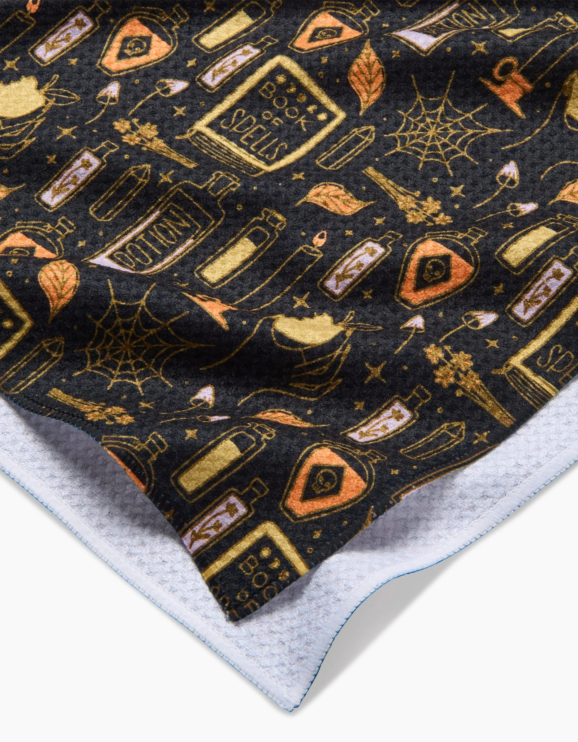 The Witchy Things tea towel by Mandy Arroyo features a Halloween-themed pattern with potion bottles, spell books, and more in gold and orange. This absorbent towel has a white textured lining for extra durability.