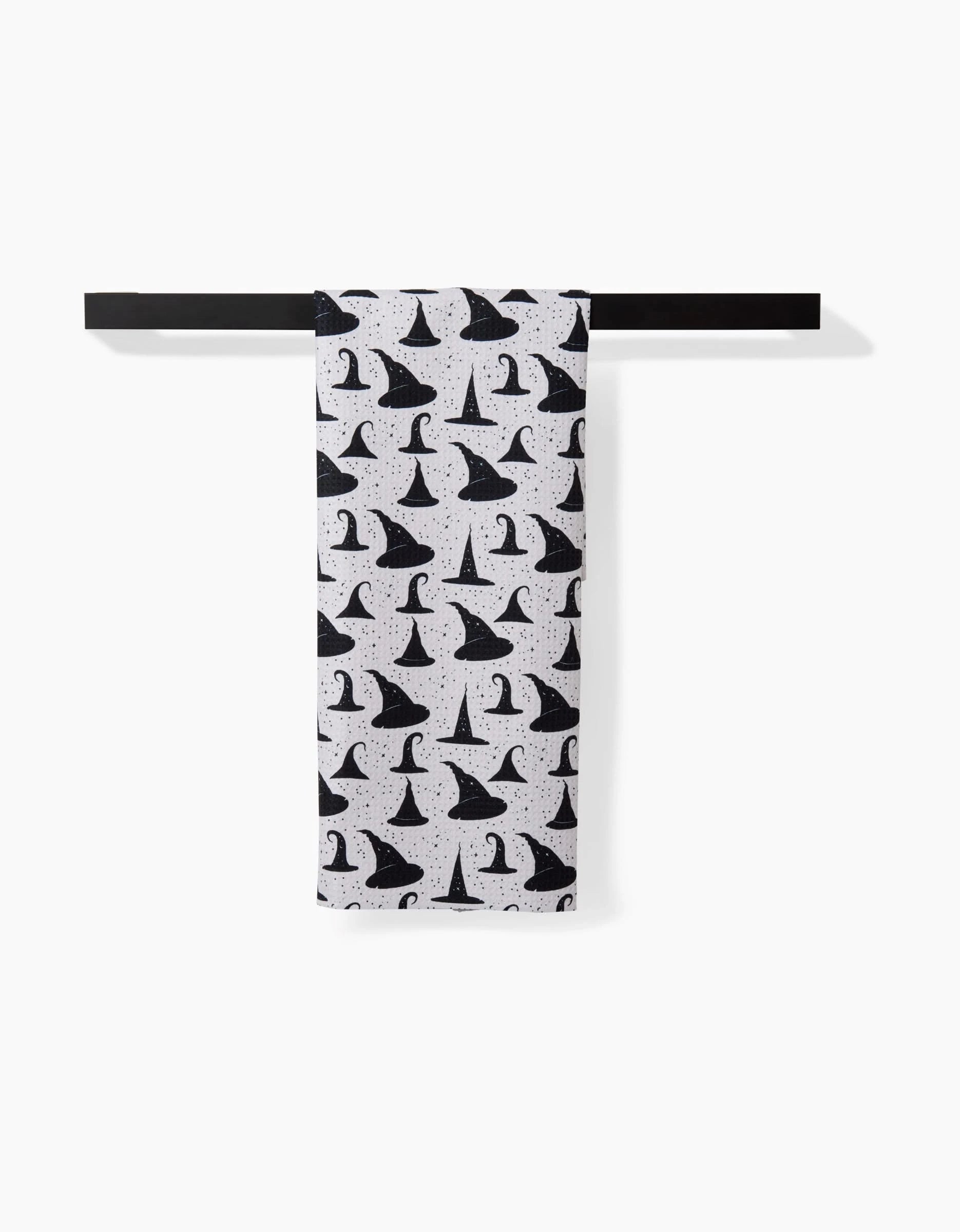 The Witchy Whims tea towel by Danelys Sidron features a black witch hat pattern on white recycled fabric, shown neatly folded over a black bar against a white background.
