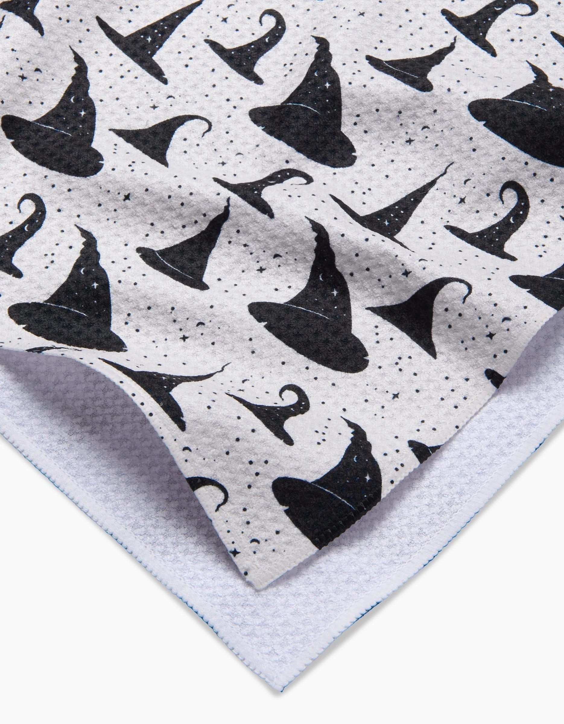 The Witchy Whims kitchen towel by Danelys Sidron is white with a textured finish and features a pattern of black witch hats and stars, shown with one corner folded to display the reverse side.