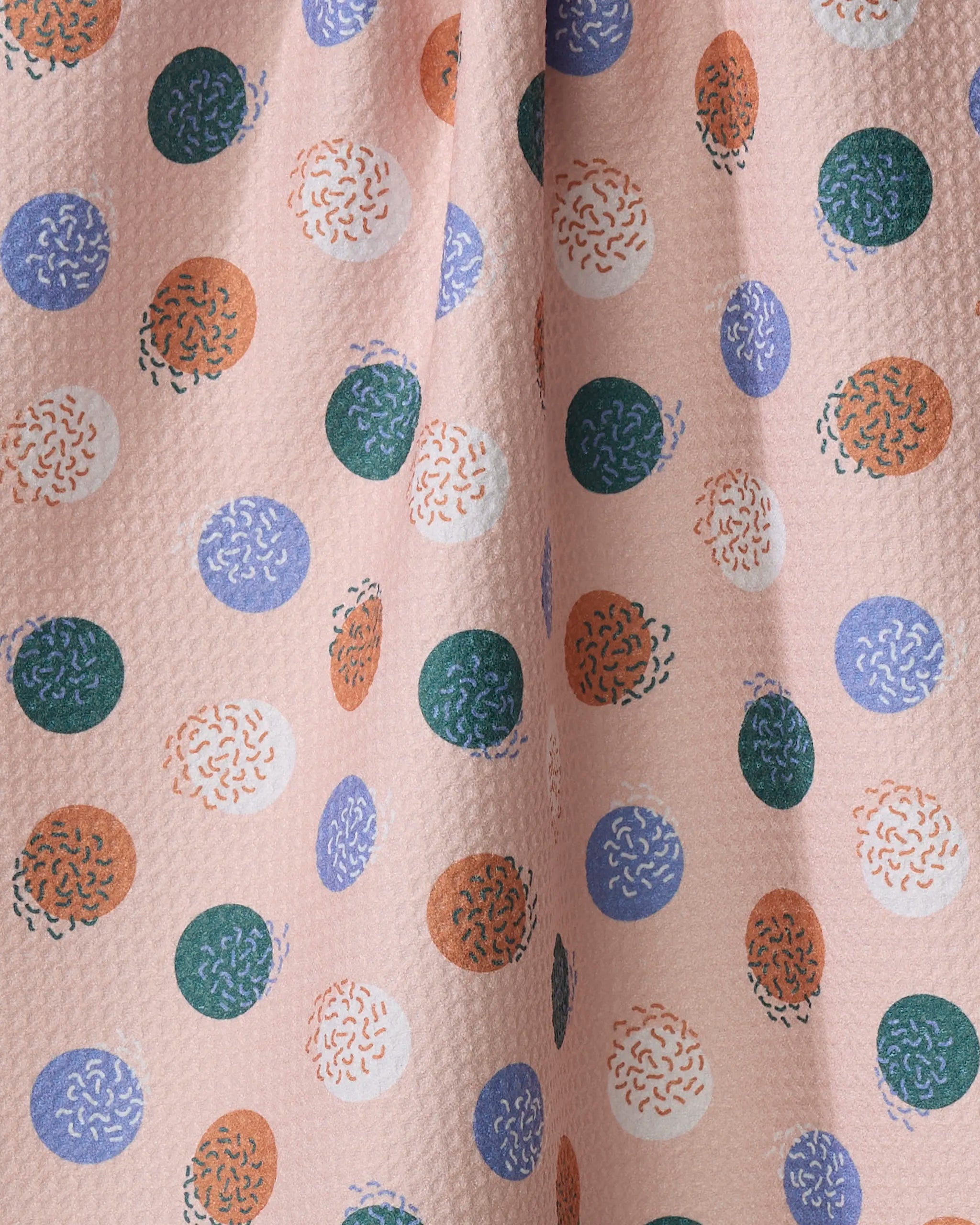 The Claire Iglesias "Wonky Dots" tea towel features a pink, slightly pleated fabric with playful green, orange, and blue circles—some solid, others squiggly—adding a vibrant touch to any kitchen.