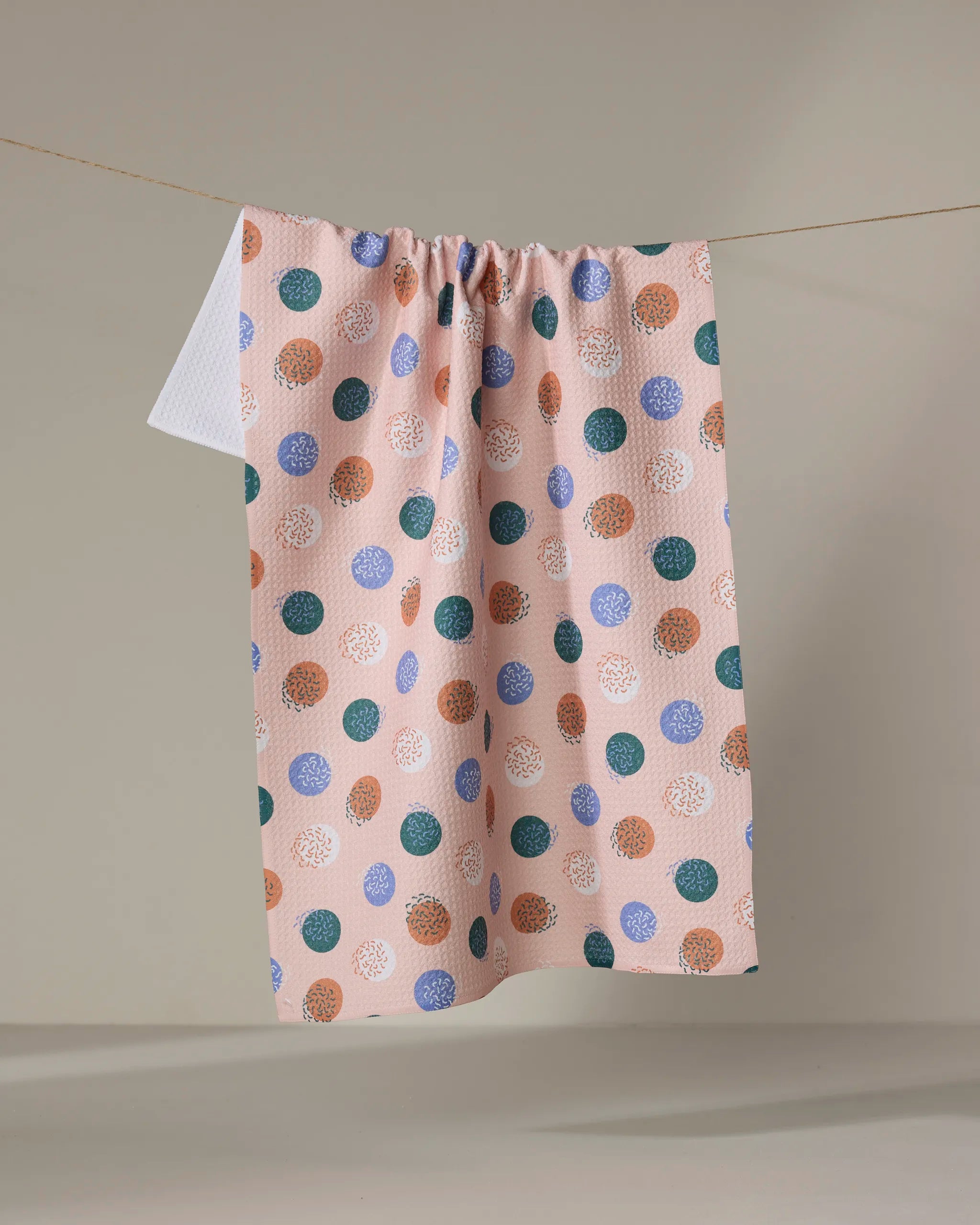 The Wonky Dots tea towel by Claire Iglesias features large blue, brown, and white polka dots on a pink background. Made from recycled materials, this eco-friendly towel brings style to any kitchen.