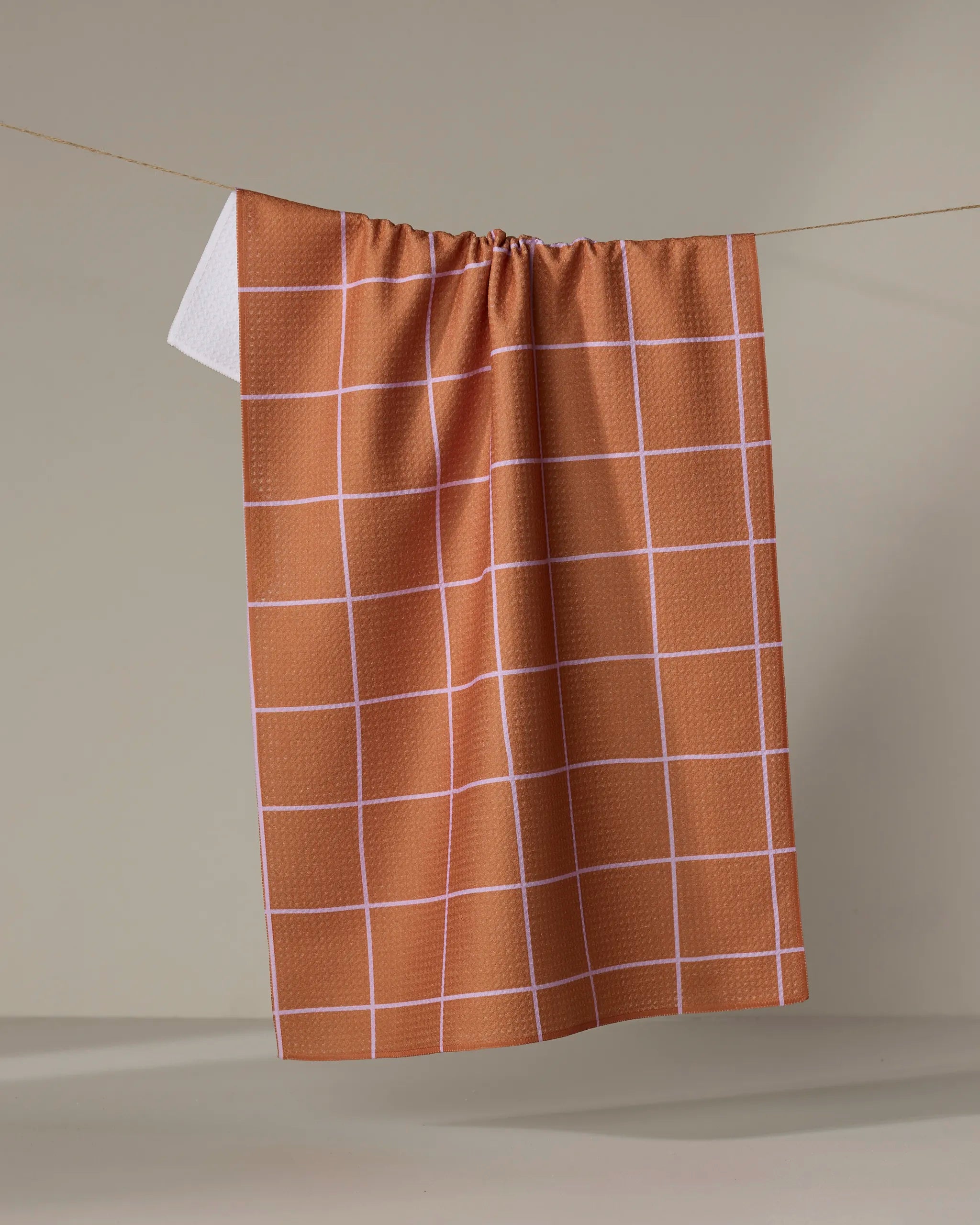 The Wonky Grid towel by Beetle Ink Co., featuring a brown color and white grid pattern, hangs on a clothesline against a plain beige background.