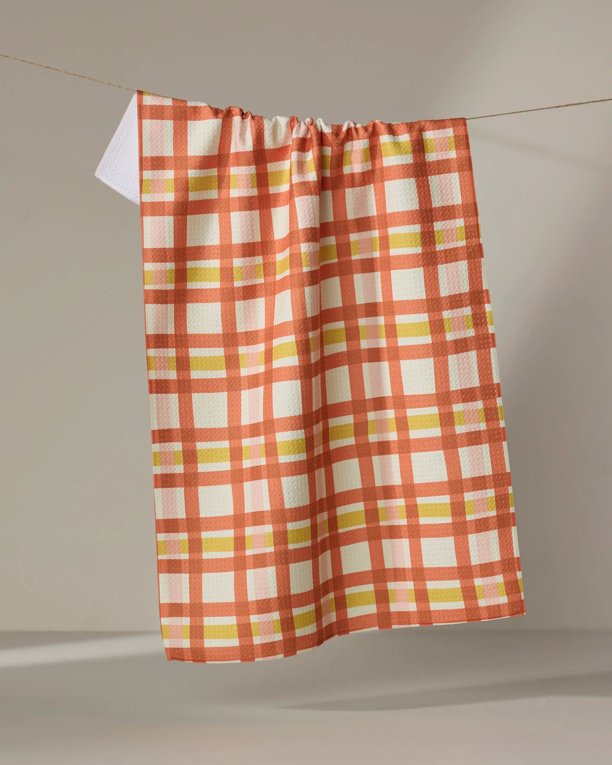 The Wonky Plaid kitchen towel by Kelsey Weigl, featuring an orange, yellow, and white plaid design, hangs on a clothesline against a neutral background.