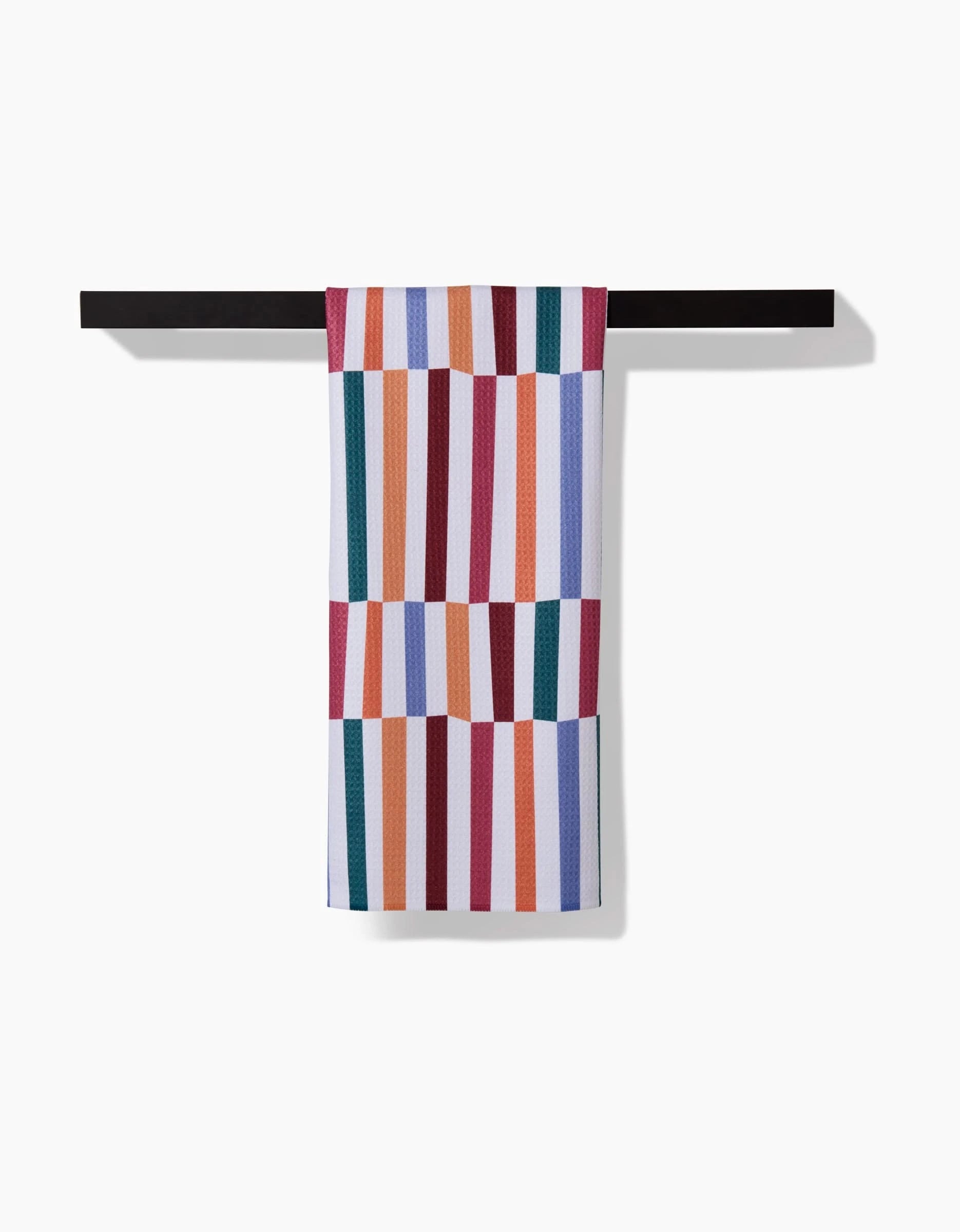 The Wonky Stripes tea towel by Claire Iglesias, made from recycled materials, features vibrant red, orange, purple, blue, and green stripes and hangs neatly over a black bar against a white background.