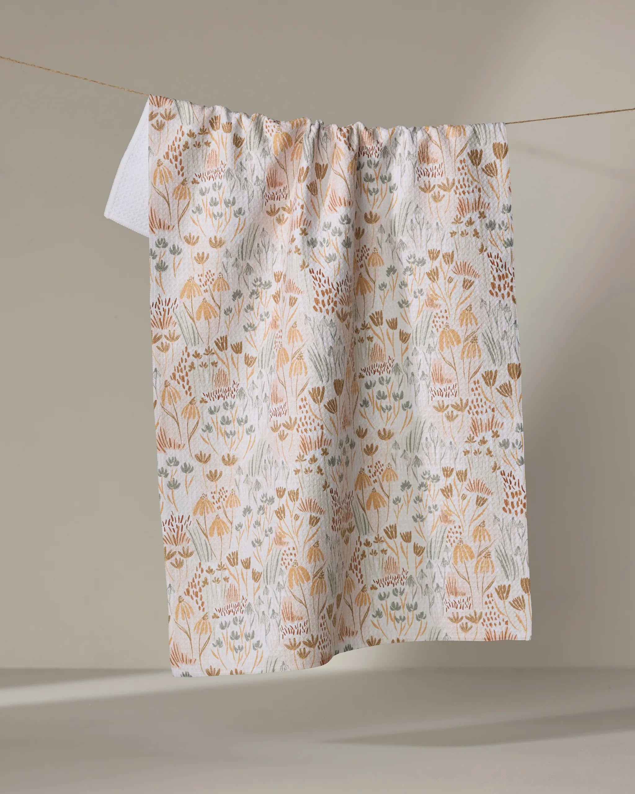 The Woodland Floral tea towel by Hufton Studio, featuring a soft beige, orange, and blue floral pattern, hangs on a clothesline against a neutral background.