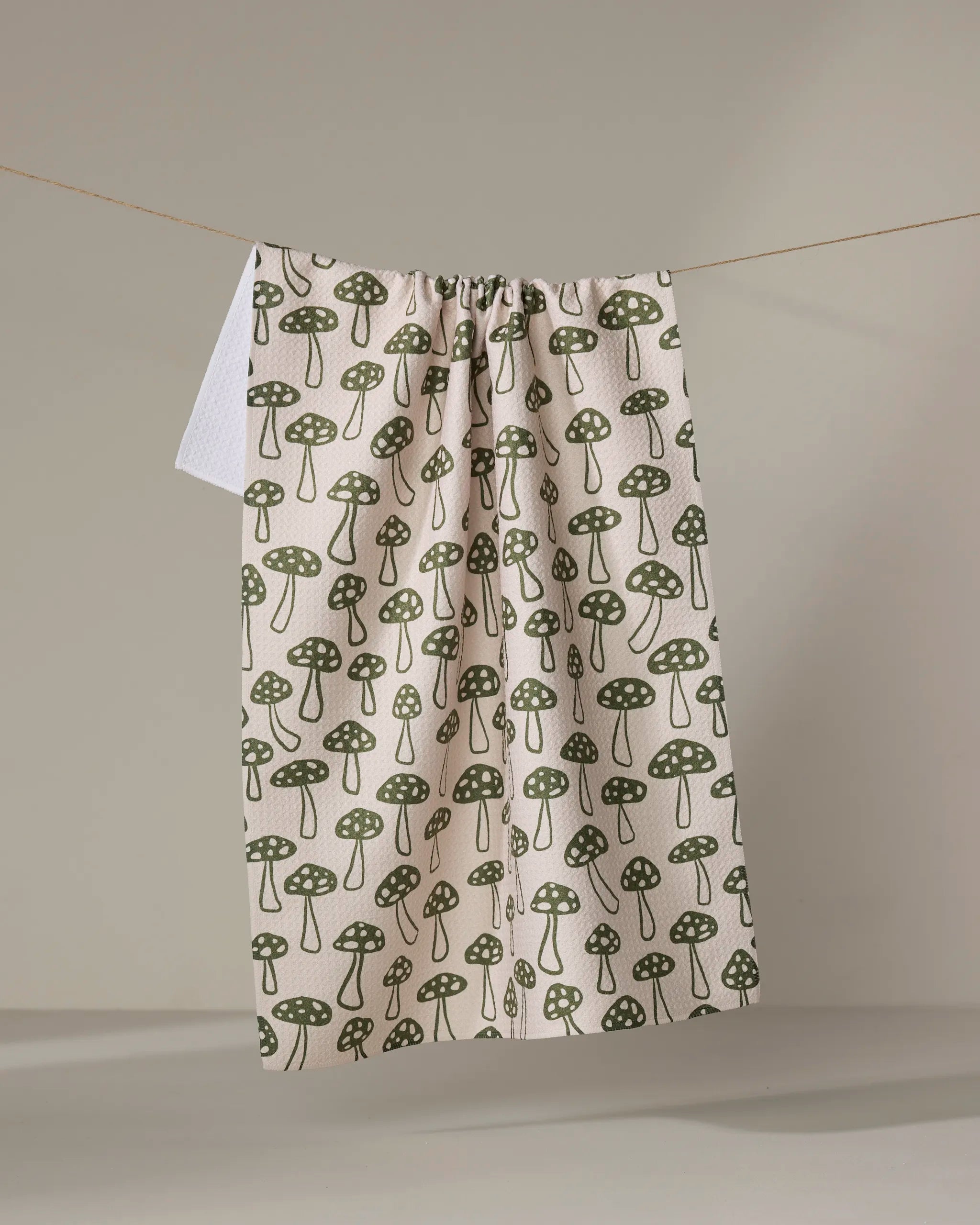 The Kayla Ann Woodland Mushroom recycled kitchen towel, featuring green mushrooms on a cream background, hangs on a string against a neutral wall.
