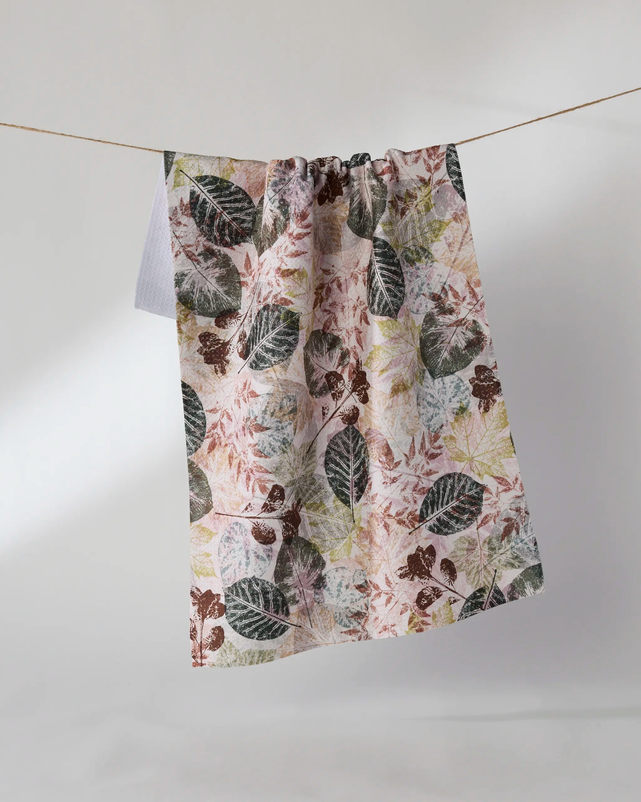 The Geometry Woodland Press towel, featuring green, brown, and pink leaf patterns, hangs on a clothesline against a plain light background.