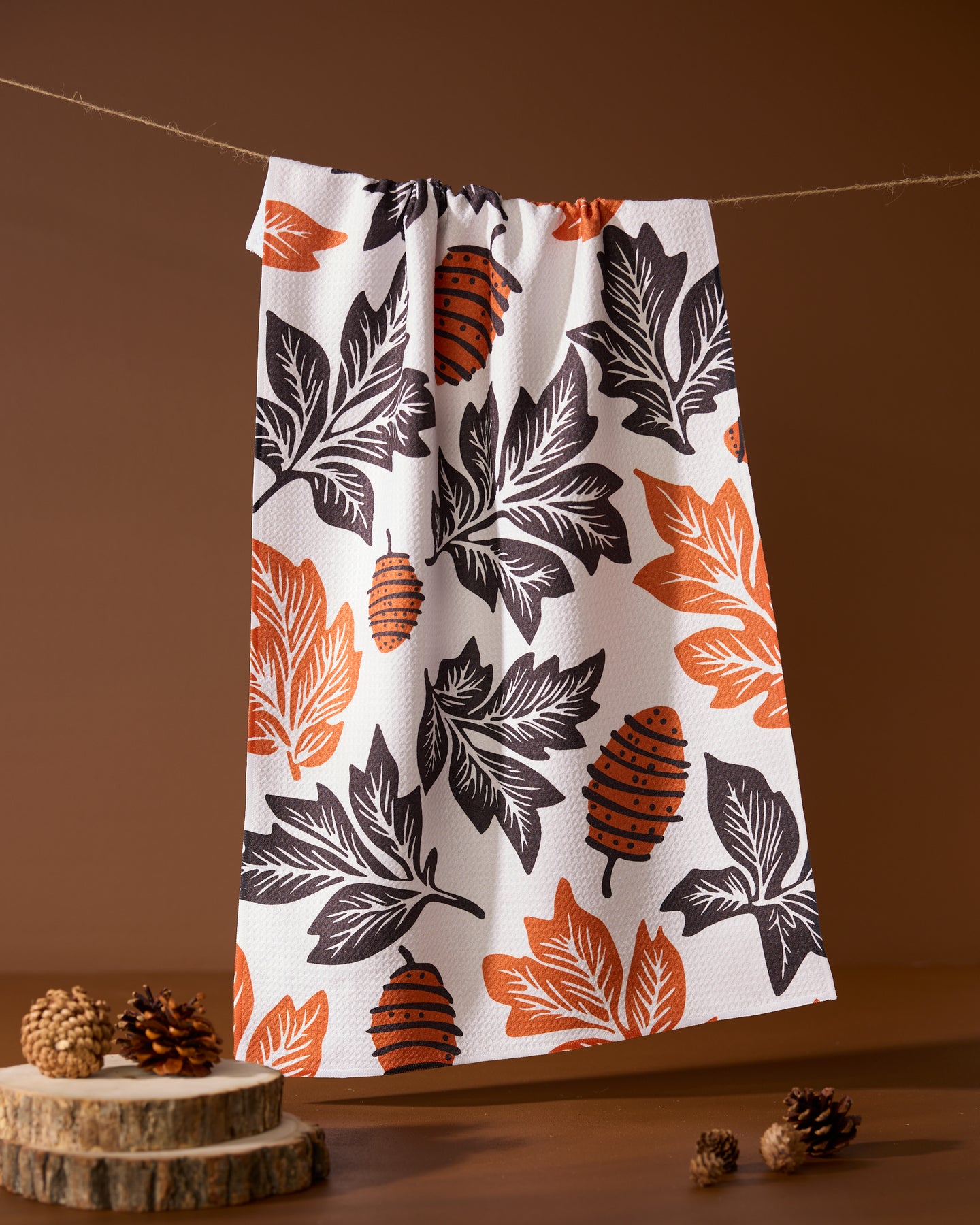 The Geometry Woodland Waltz kitchen tea towel, made from recycled materials, showcases orange and black leaf and pinecone patterns. It hangs above a brown background with pinecones and wooden slices arranged below.