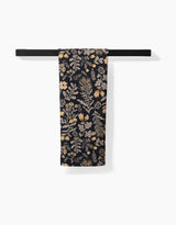 The Danelys Sidron Woodland Wildflowers tea towel, featuring a beige and gold botanical pattern on dark fabric made from recycled materials, hangs folded over a black towel bar against a white background.