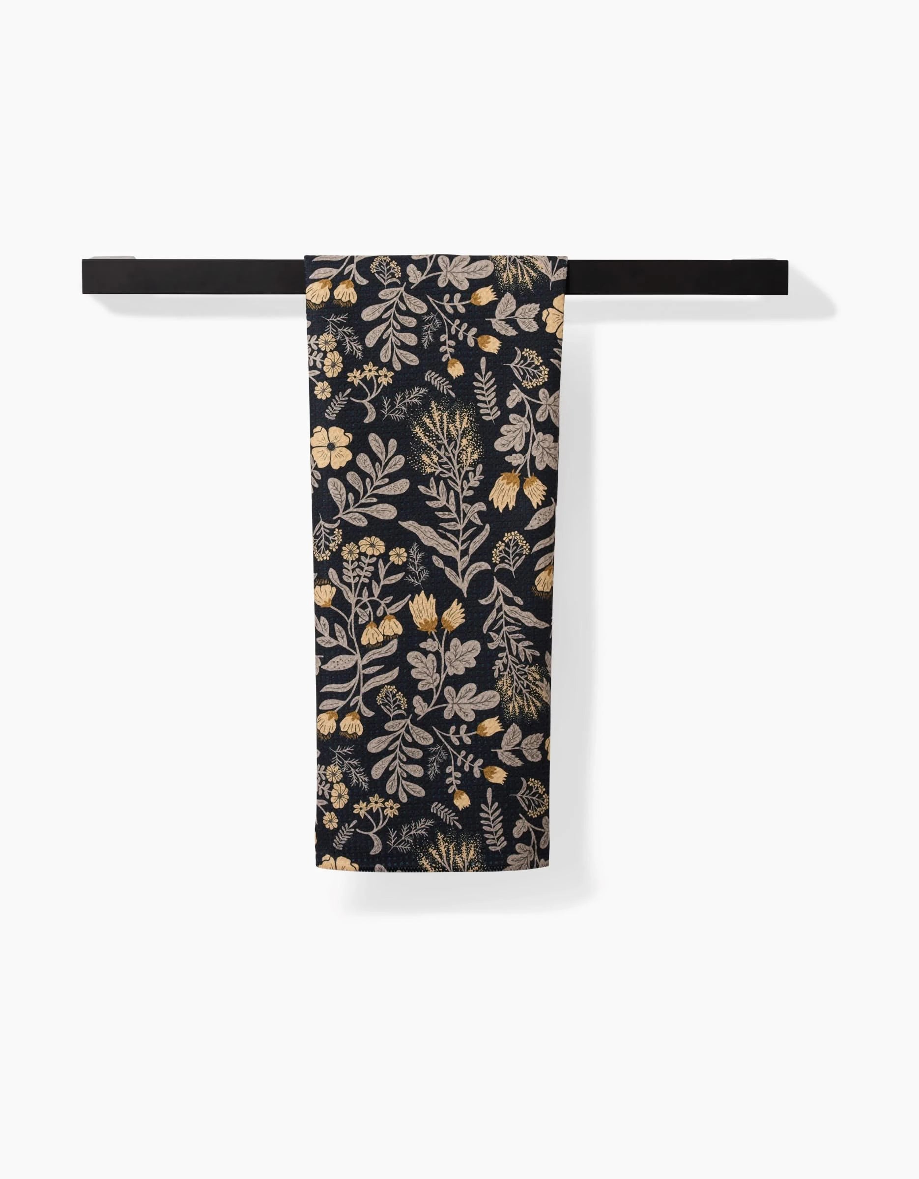 The Danelys Sidron Woodland Wildflowers tea towel, featuring a beige and gold botanical pattern on dark fabric made from recycled materials, hangs folded over a black towel bar against a white background.
