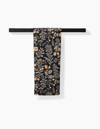 The Danelys Sidron Woodland Wildflowers tea towel, featuring a beige and gold botanical pattern on dark fabric made from recycled materials, hangs folded over a black towel bar against a white background.