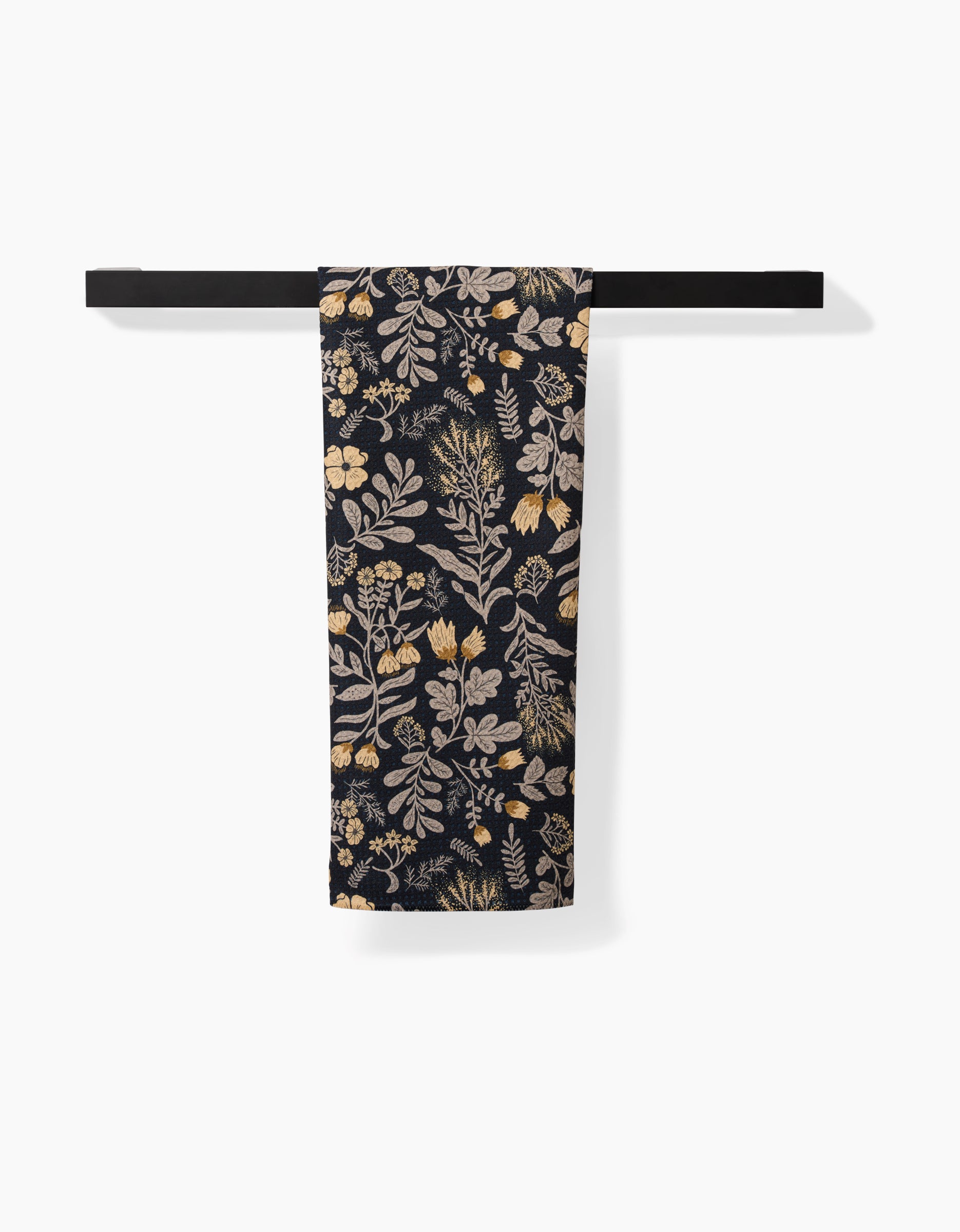 The Danelys Sidron Woodland Wildflowers tea towel, featuring a beige and gold botanical pattern on dark fabric made from recycled materials, hangs folded over a black towel bar against a white background.